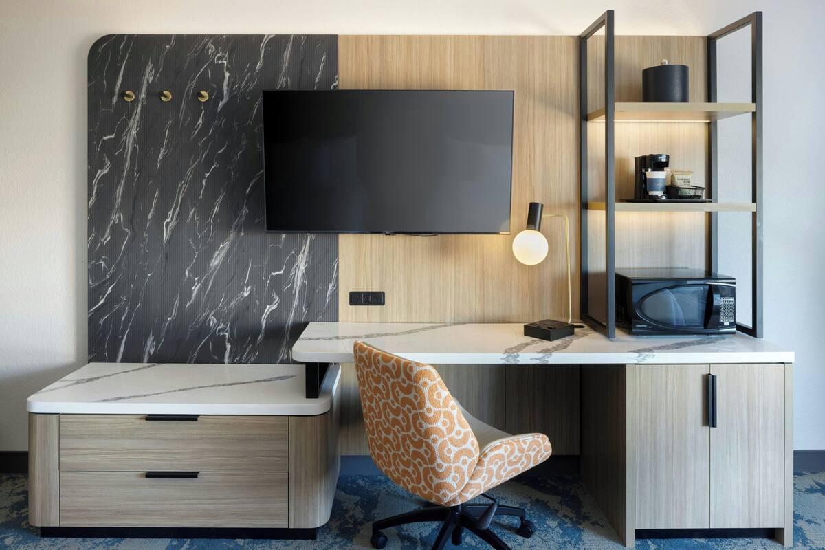 Een moderne hotelkamer met een flatscreen televisie bevestigd aan een marmeren muur, een bureau met stoel, een koffieapparaat en een magnetron op een plank.