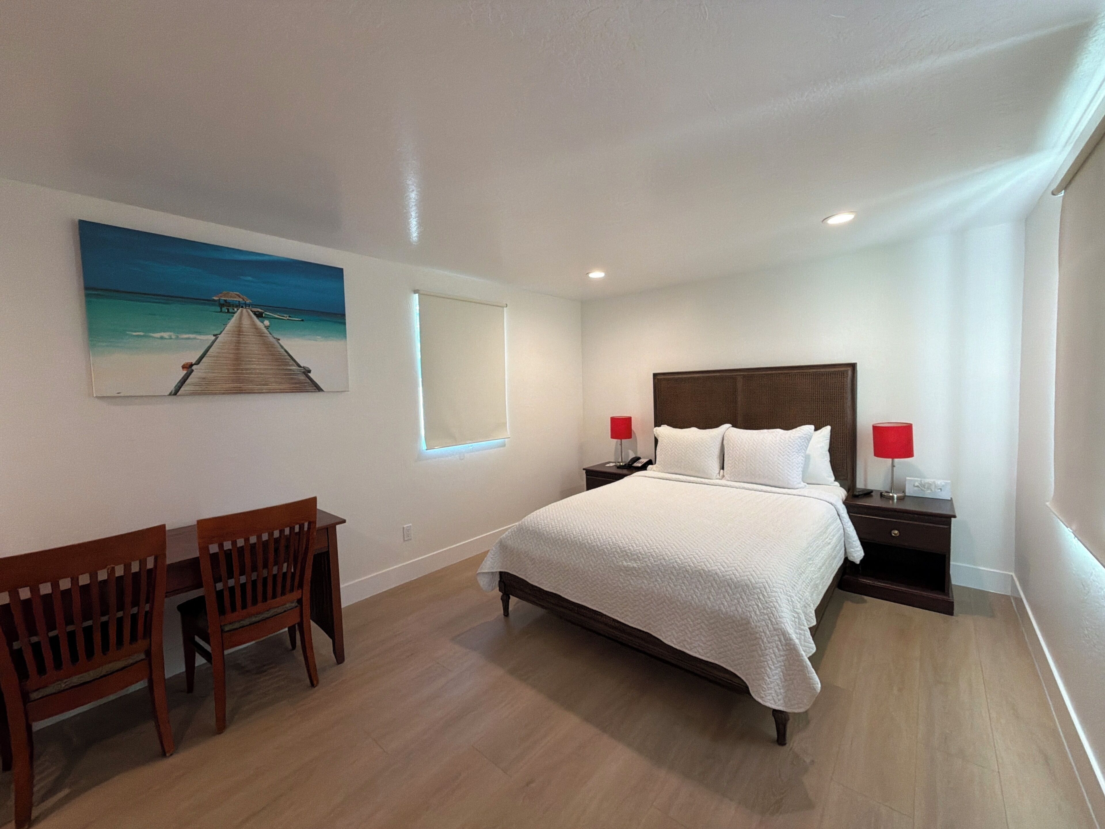 Une chambre d’hôtel avec un lit, deux tables de chevet, un bureau avec des chaises, un tableau au mur et une fenêtre.