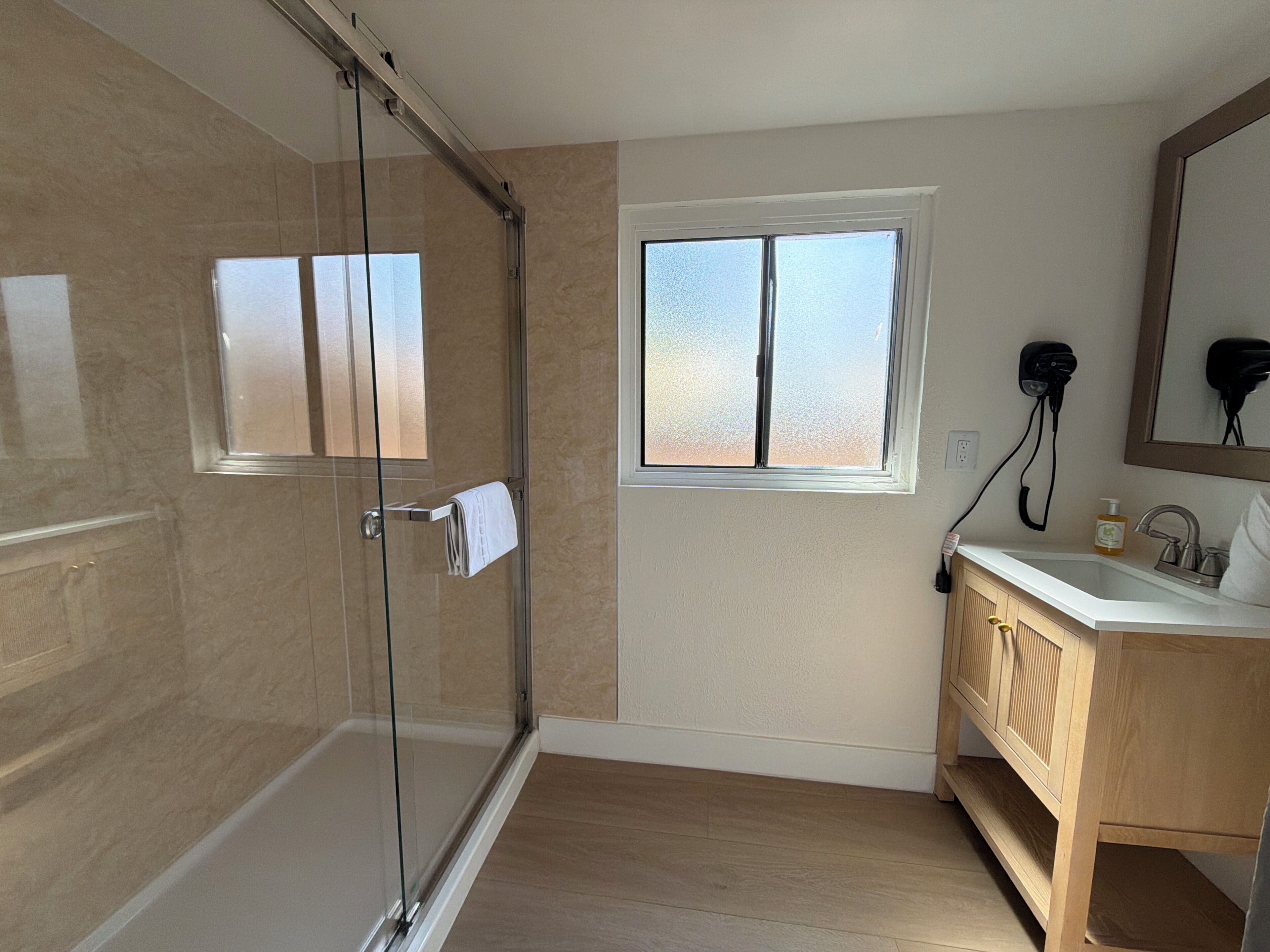 A bathroom with a glass shower door, a window, a sink with a mirror, and wooden cabinetry.