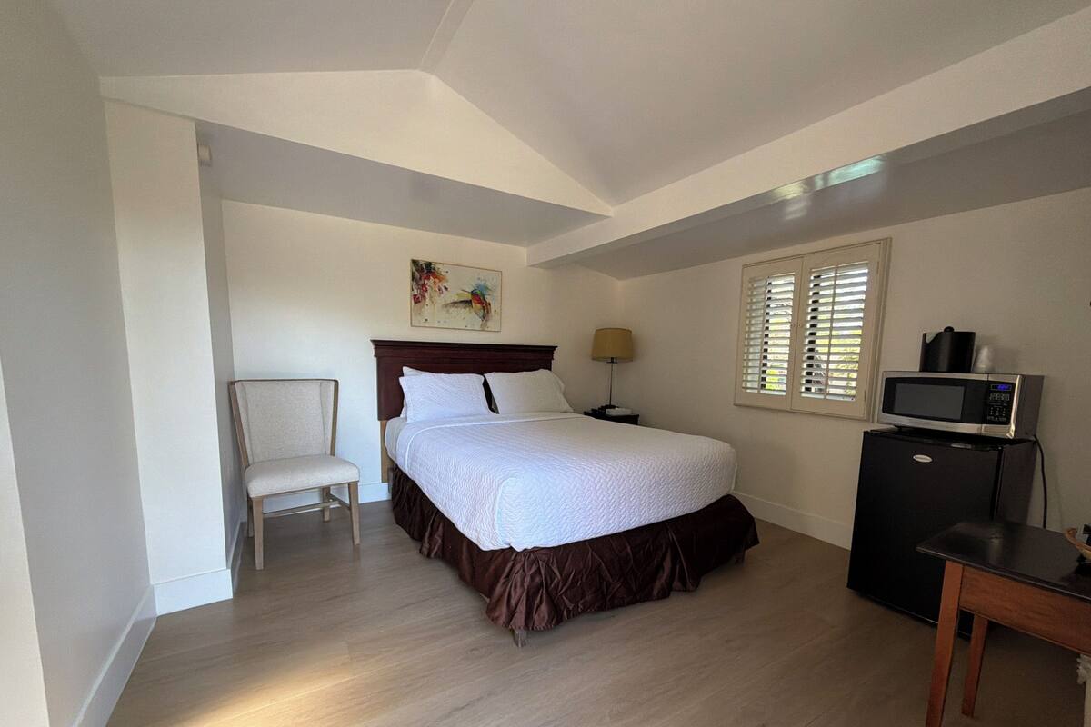 Un dormitorio con cama, una silla, un escritorio, un televisor y una ventana con persianas.