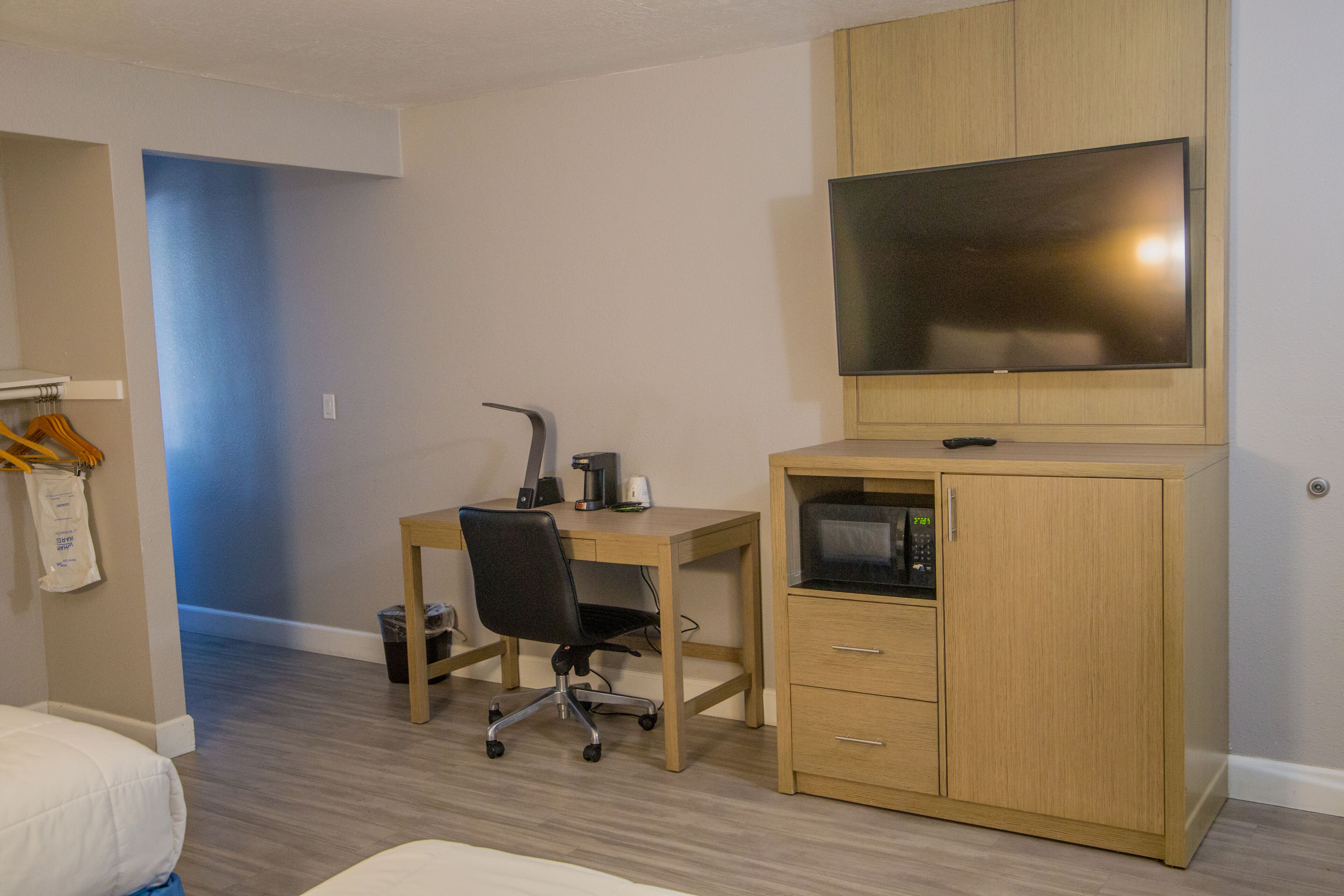A hotel room with a wooden desk, a black office chair, a television mounted on the wall, and a bed with white bedding.