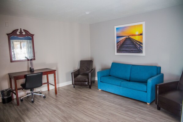Una habitación con escritorio, dos sillas, un sofá azul, piso de madera, espejo adosado a la pared y una pintura de atardecer.