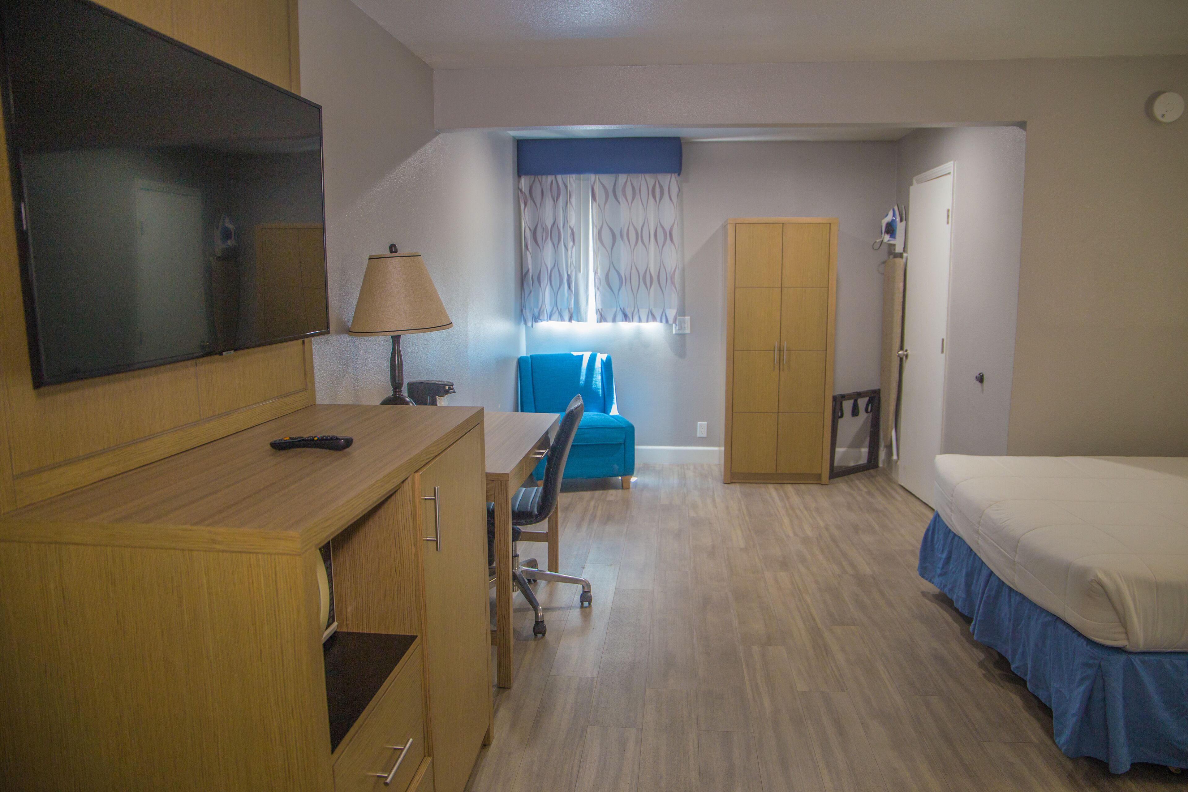 A hotel room with a wooden desk, a TV, a bed, a wardrobe, a chair, and a window with curtains.