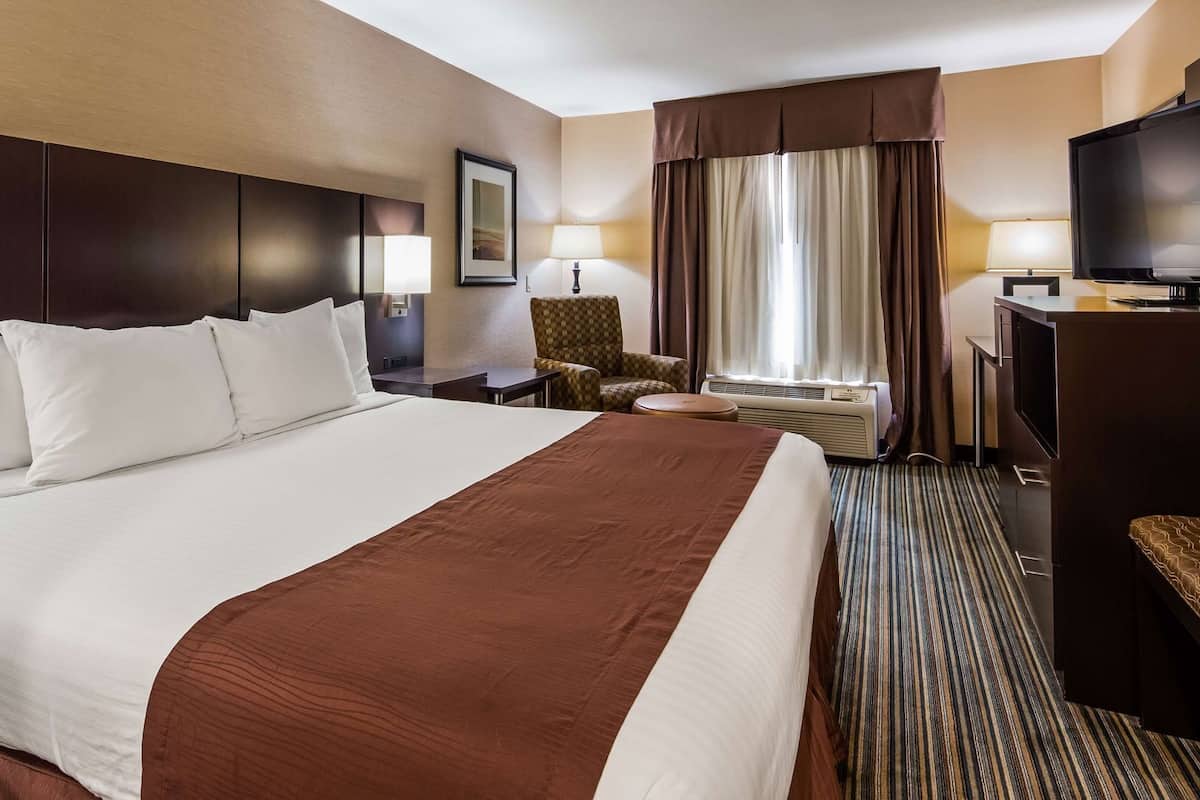 Ein Hotelzimmer mit einem großen Bett, einem Schreibtisch mit Fernseher, einem Sessel, einer Lampe und einem Fenster mit Vorhängen.