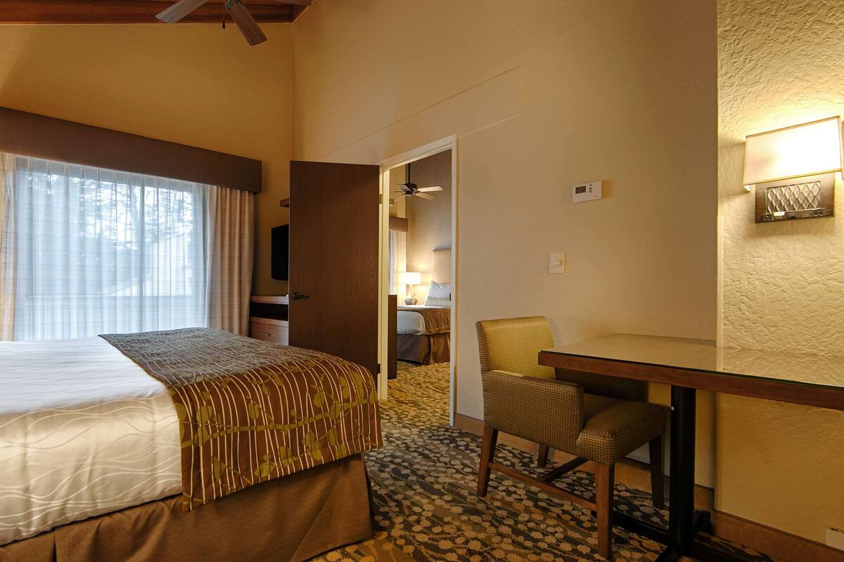 A hotel room with a bed, a desk, a chair, a window with curtains, and a door leading to another room.
