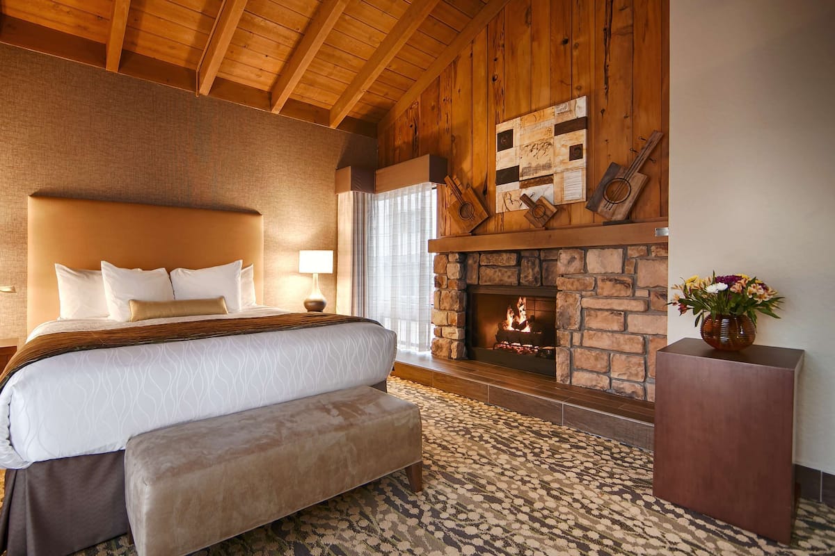 A bedroom with a large bed, wooden ceiling beams, a stone fireplace, and a patterned carpet.