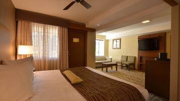 Una habitación de hotel con cama, ventilador de techo, televisión y zona de estar con sillas y mesa.