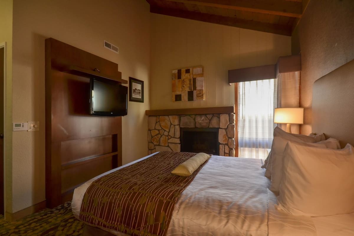 A hotel room with a bed, a television mounted on the wall, a stone fireplace, and a window with curtains.