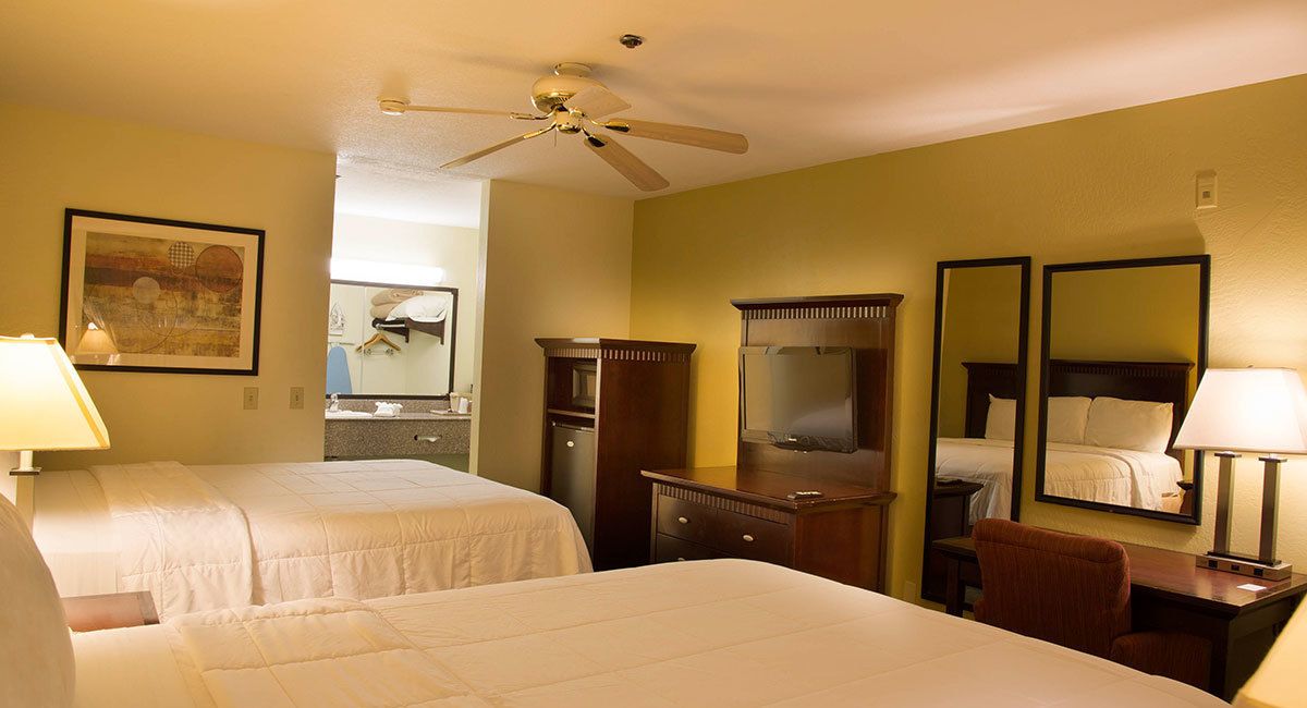A hotel room with two beds, a desk, a chair, a wardrobe, a television, and a ceiling fan.