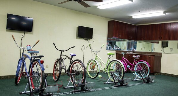 A room with four stationary bikes, a ceiling fan, and two wall-mounted televisions.