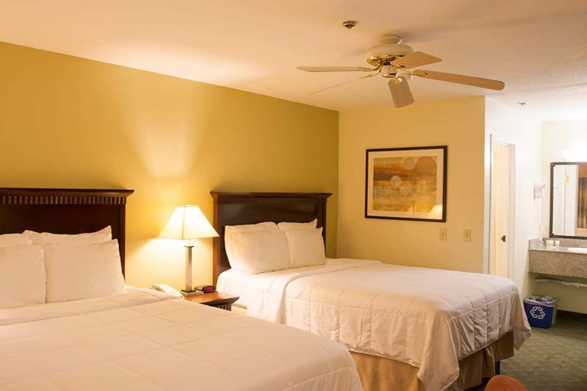 A hotel room with two beds, a ceiling fan, a bedside lamp, a framed picture on the wall, and a bathroom visible through an open door.
