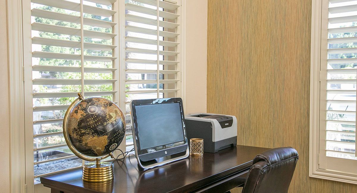 A desk with a globe, a laptop, and a printer in a room with wooden paneling and windows with blinds.