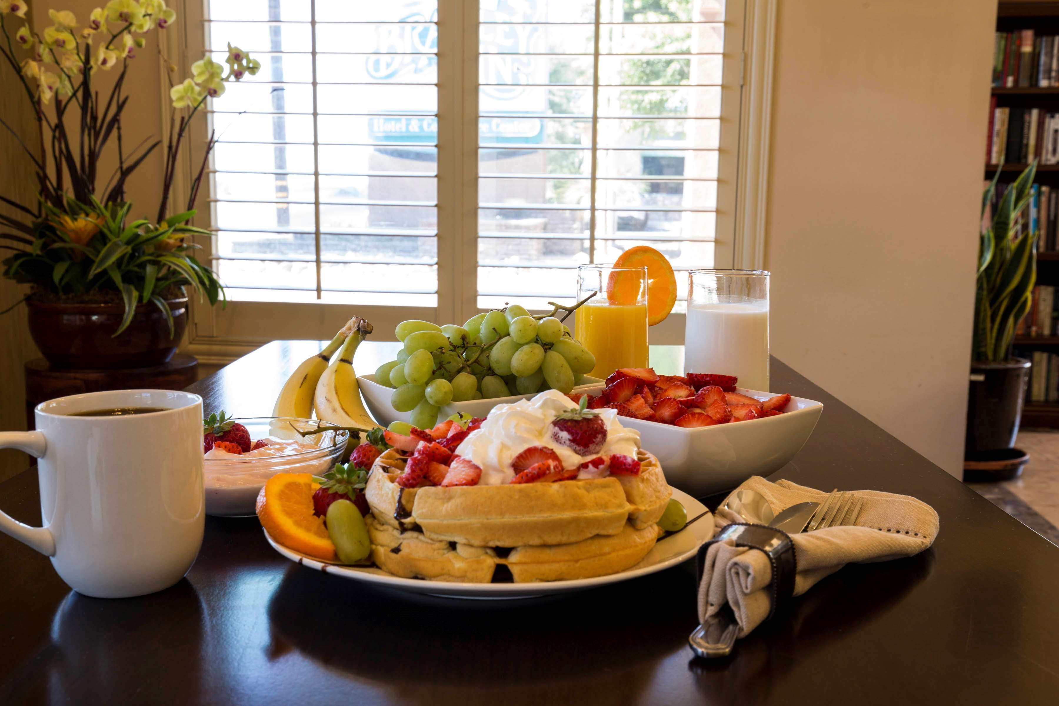 Une table de petit-déjeuner avec une variété de fruits, une assiette de gaufres garnies de crème fouettée et de fraises, un verre de jus d’orange et une tasse de café.