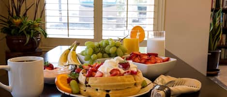 A breakfast table with a variety of fruits, a plate of waffles topped with whipped cream and strawberries, a glass of orange juice, and a cup of coffee.