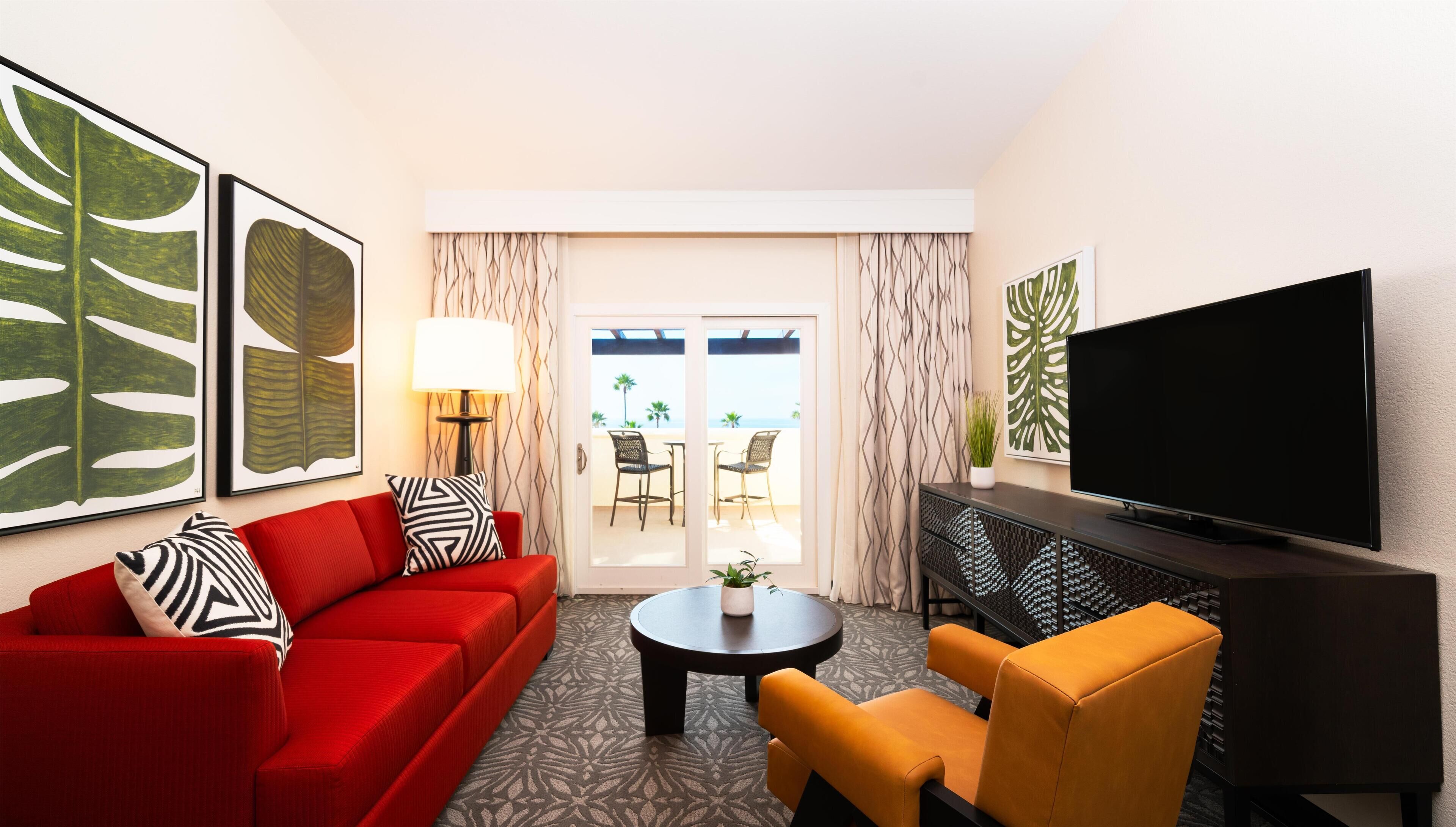 A modern living room with a red sofa, a yellow armchair, a flat-screen TV, and a large window with a view of palm trees.