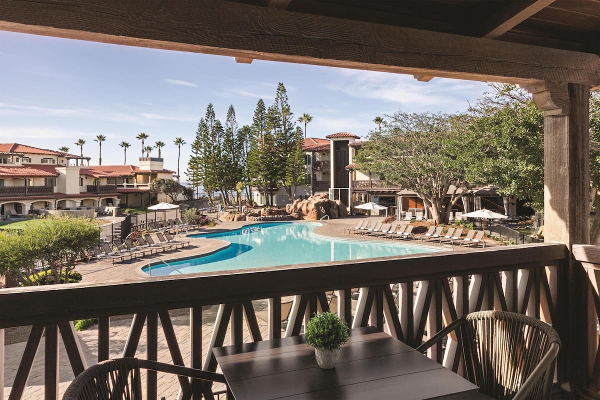 A poolside area with lounge chairs and a table, overlooking a swimming pool and a landscaped garden with trees and a building in the background.