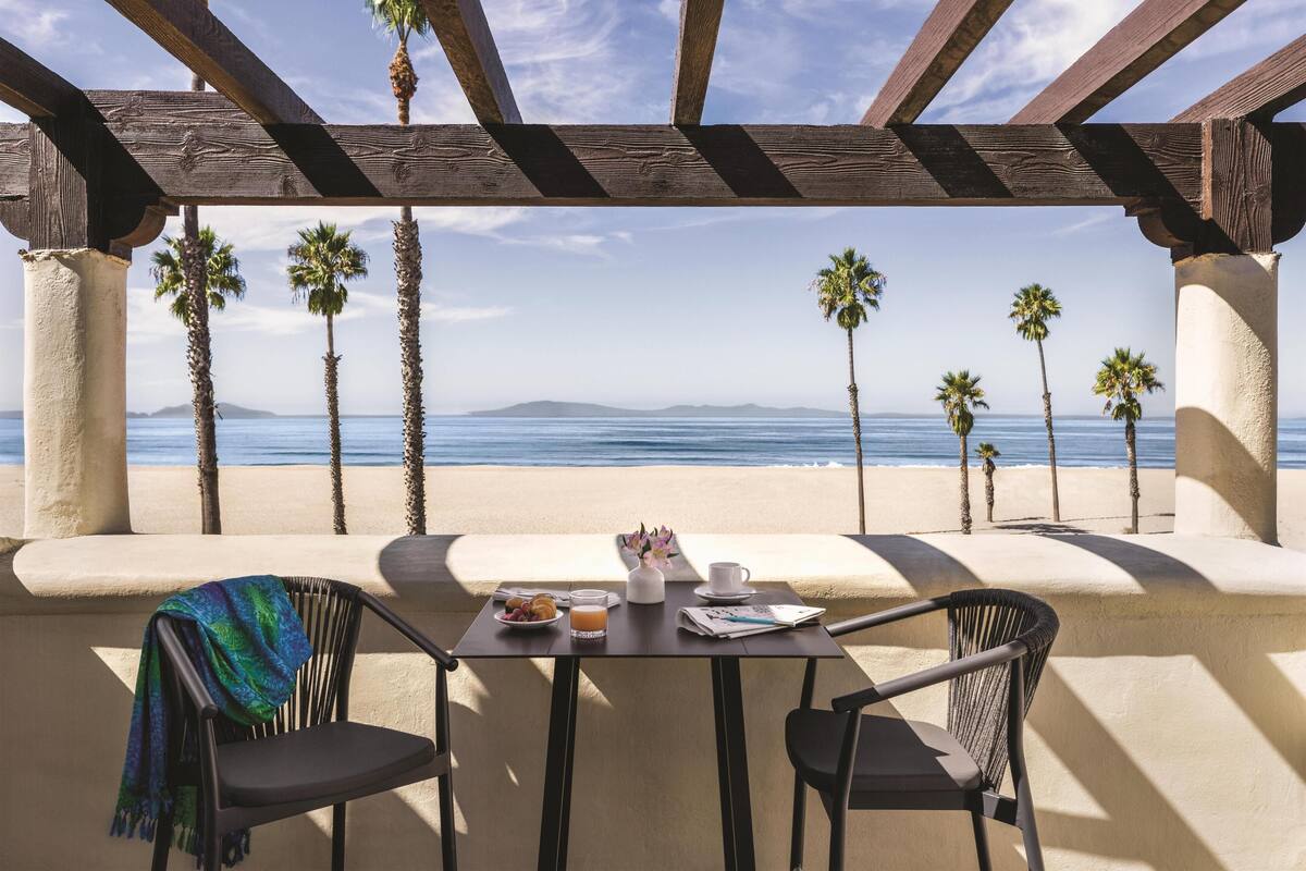 A table with a view of the beach and ocean, a chair with a blanket, and a small table with a cup, plate, and glass.