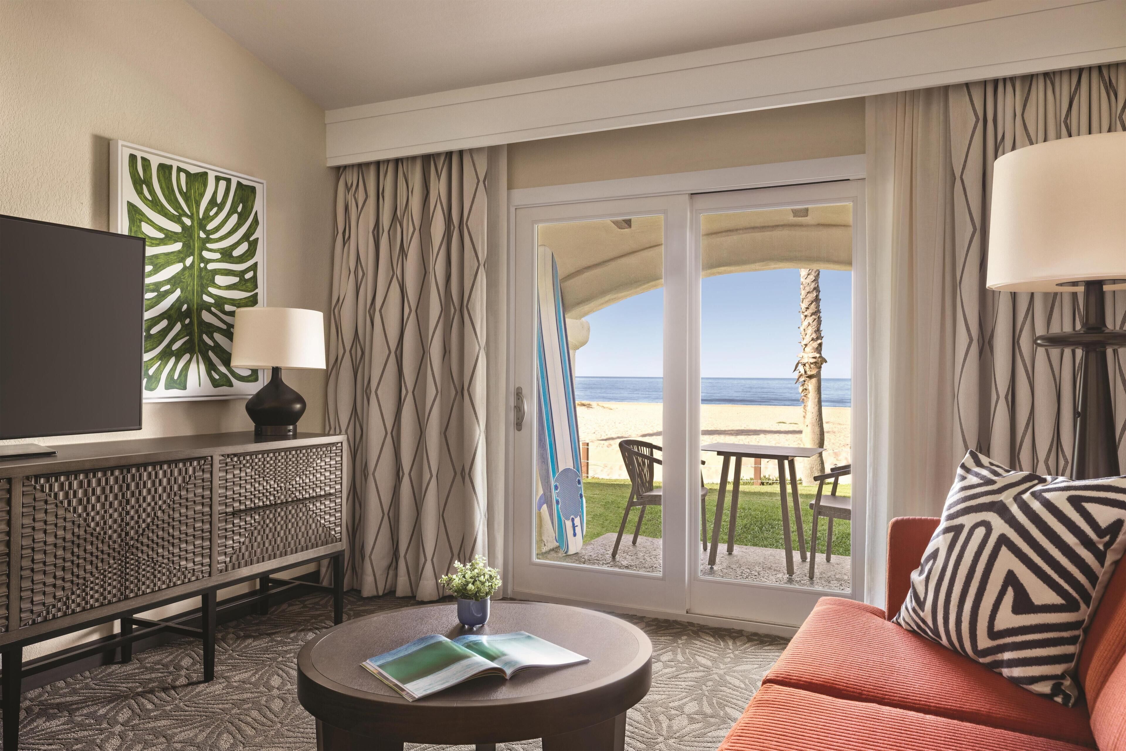 Een hotelkamer met een televisie, een bank, een salontafel, uitzicht op het strand en een balkon met een tafel en stoelen.