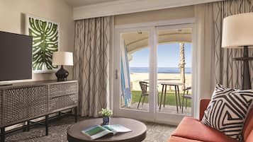 Een hotelkamer met een televisie, een bank, een salontafel, uitzicht op het strand en een balkon met een tafel en stoelen.
