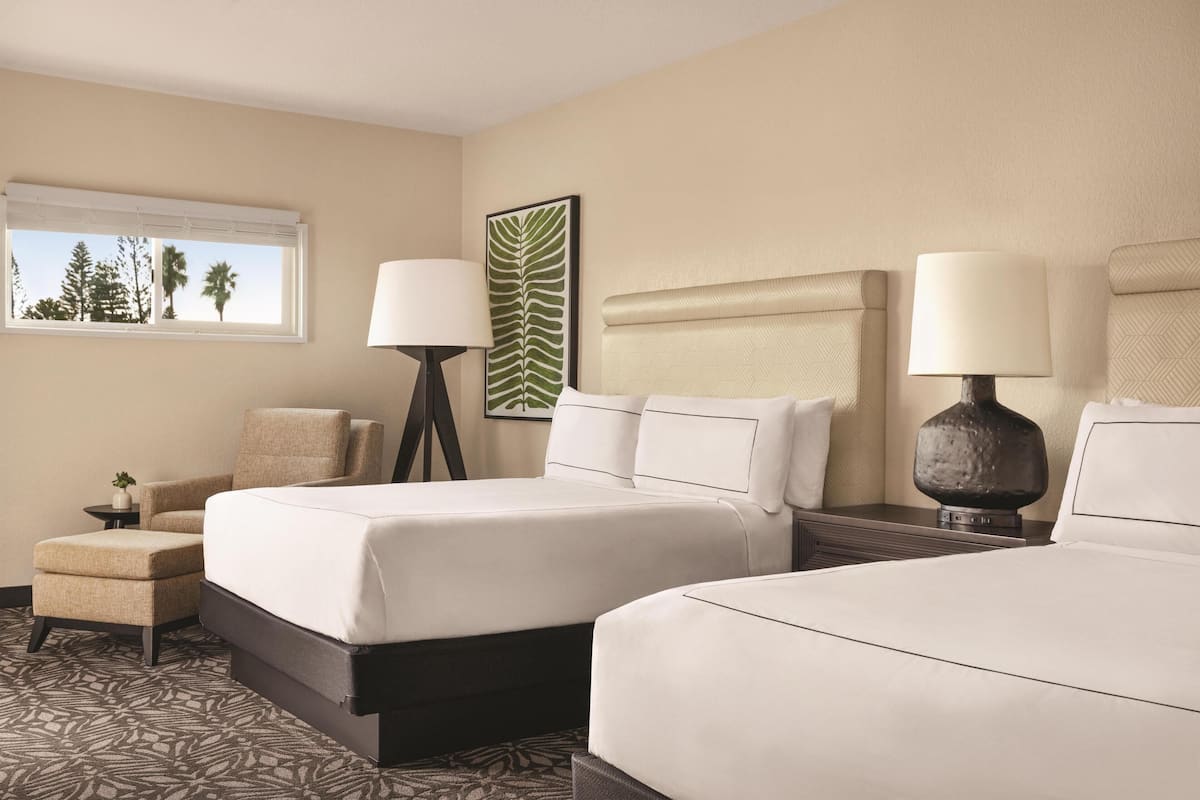 A hotel room with two beds, a chair, a small table with a lamp, and a window with a view of palm trees.