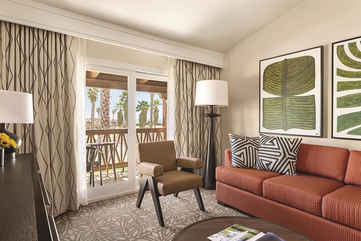 A hotel room with a sofa, armchair, and a large window with a view of palm trees.