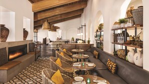 Un'elegante area lounge dal design moderno, caratterizzata da soffitti alti, travi in legno a vista e un camino. Sono presenti comode zone relax con cuscini e tavoli rotondi apparecchiati per mangiare.