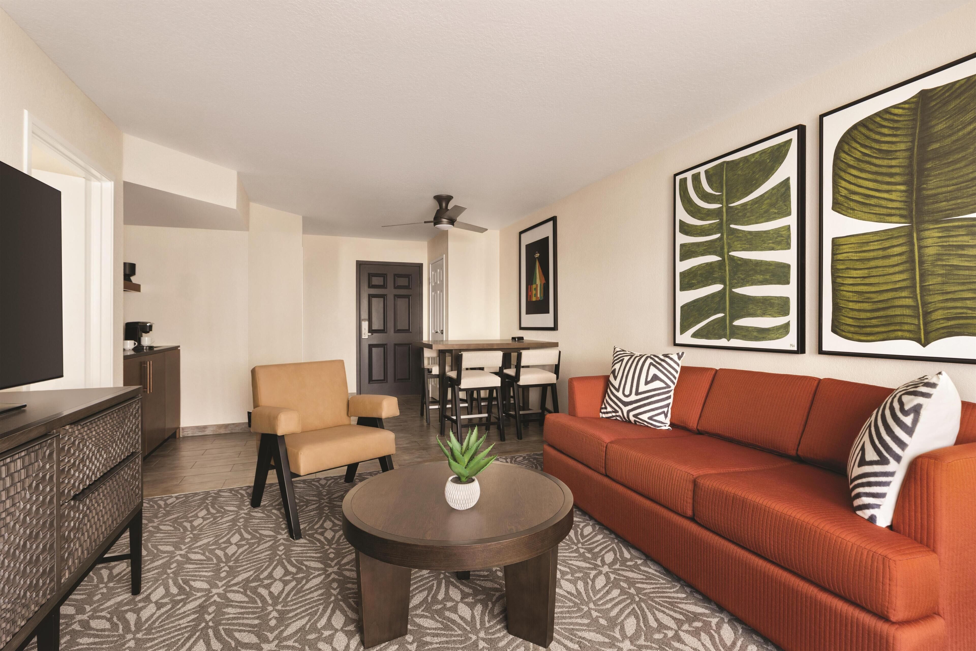 A living room with a brown sofa, a beige armchair, a coffee table with a plant, and a TV on a cabinet. There are framed leaf artworks on the wall.
