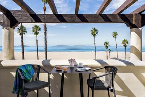 Una mesa con vista a la playa y al océano, una silla con una manta, y una mesita con una taza y un plato de comida.