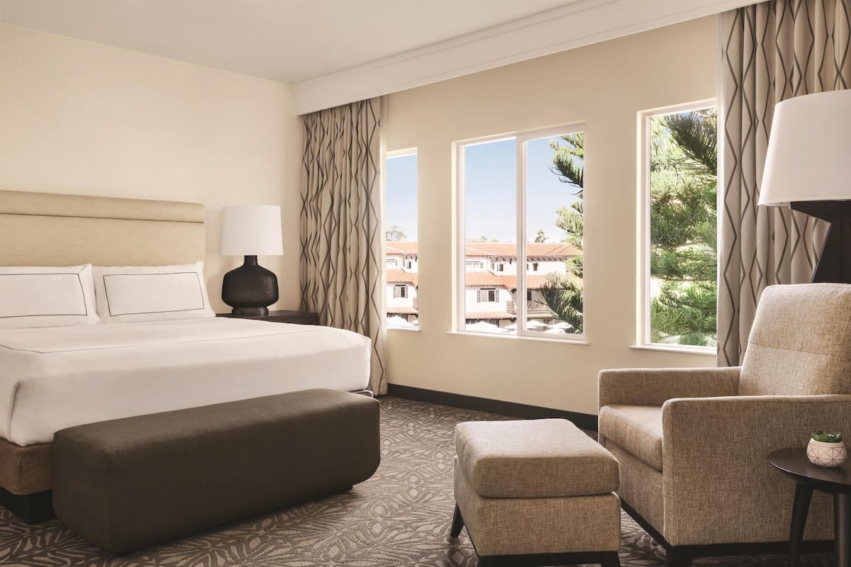 A hotel room with a large bed, a sitting area with a chair and ottoman, a lamp, and a view of buildings and greenery through the windows.