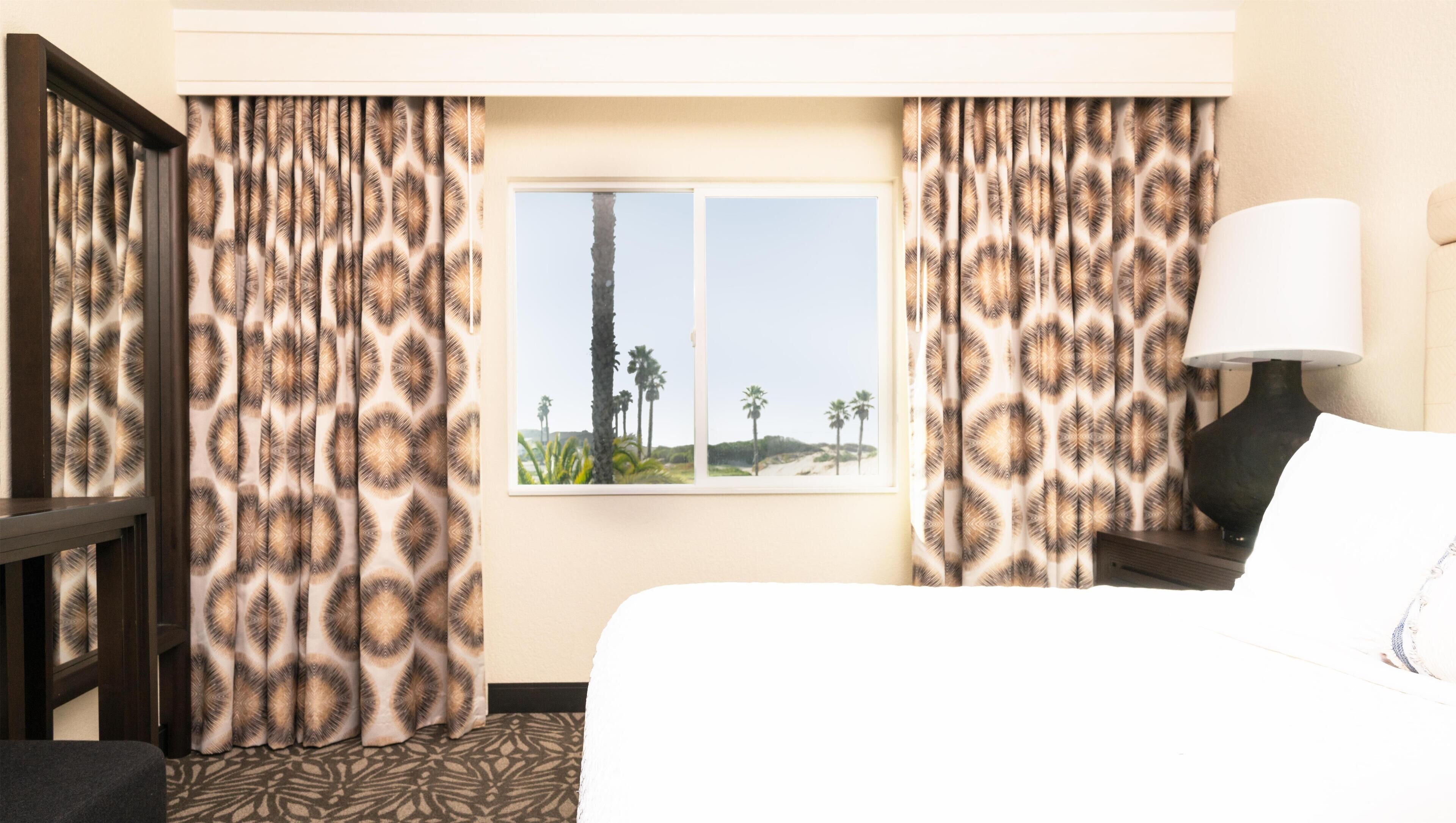 Een hotelkamer met een bed, een nachtkastje met een lamp, een raam met uitzicht op palmbomen en kamergordijnen met een patroon.