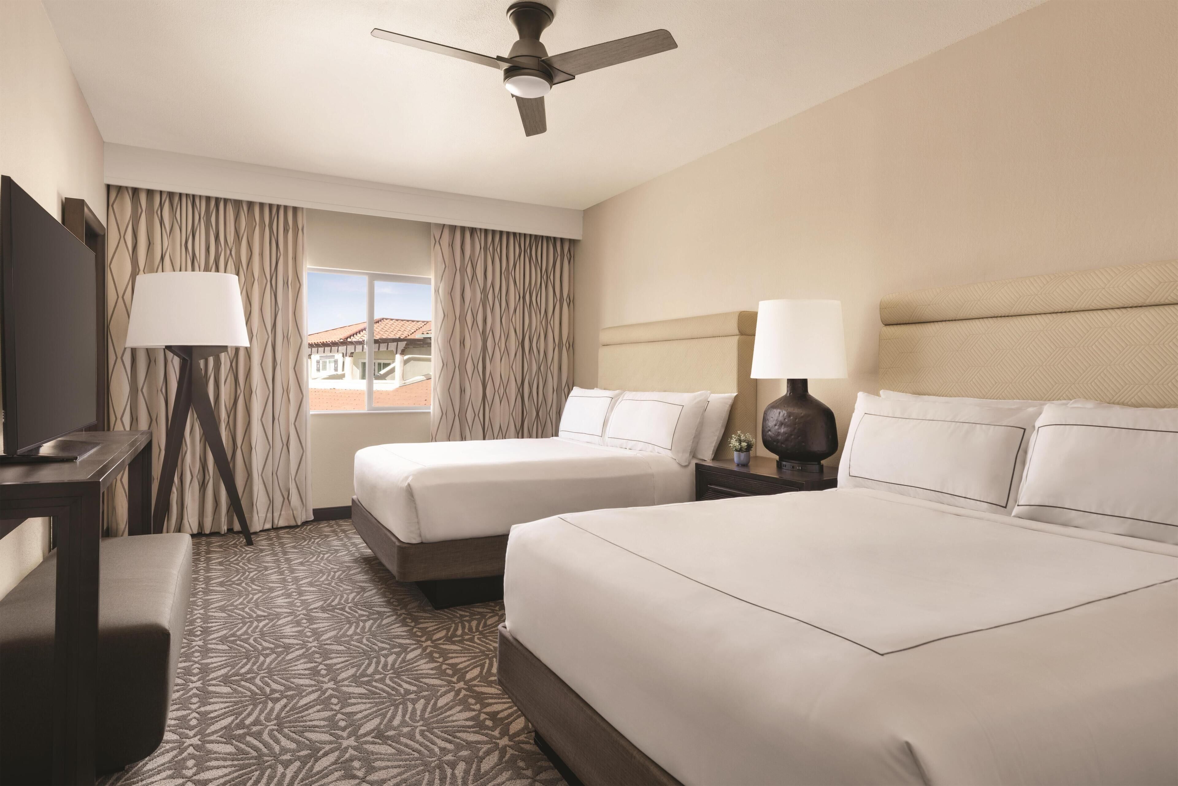 A hotel room with two beds, a desk, a chair, a lamp, and a ceiling fan.