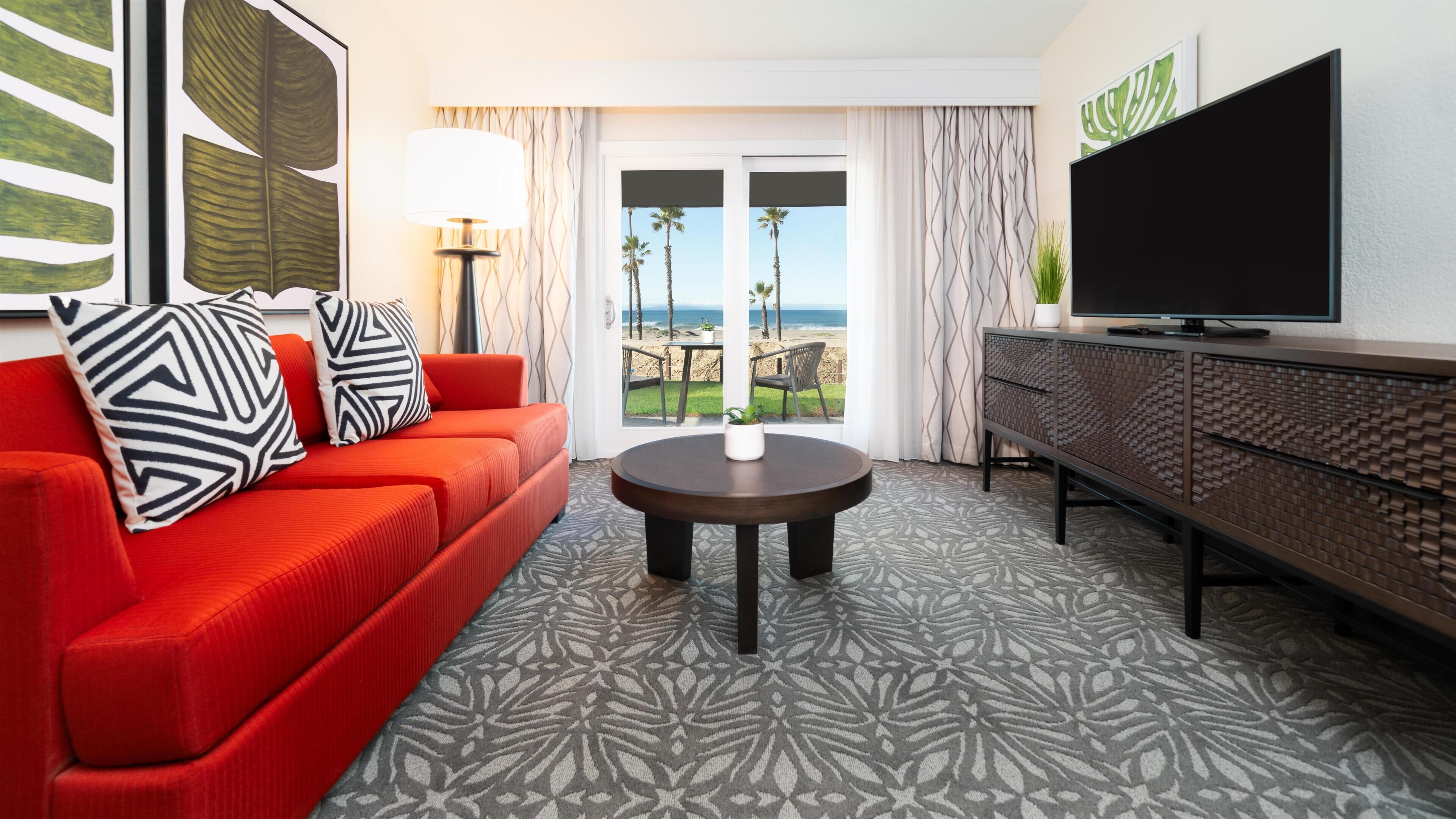 Een hotelkamer met een rode bank, een ronde tafel, een televisie en uitzicht op het strand.
