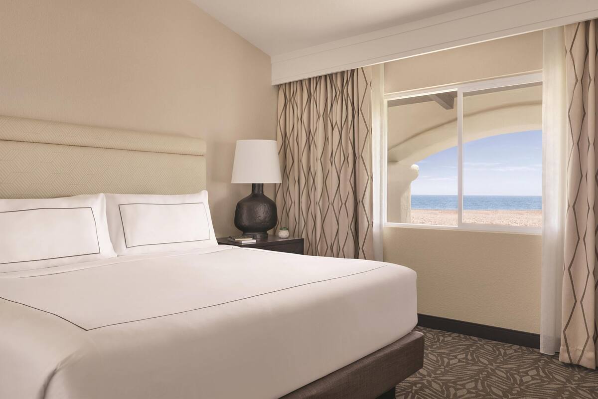 A hotel room with a bed, a nightstand with a lamp, and a view of the beach.