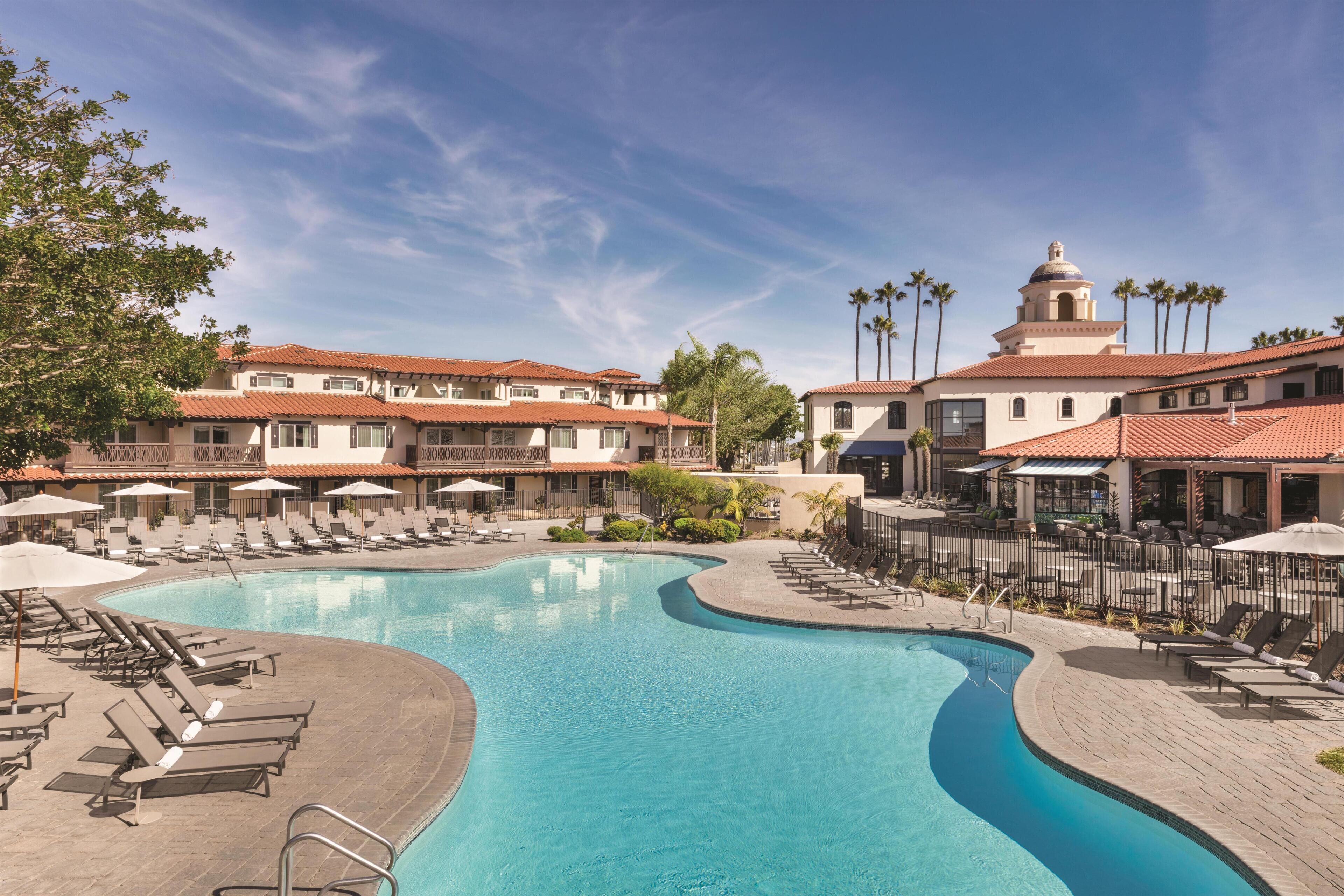 A swimming pool with lounge chairs and umbrellas, surrounded by buildings with red-tiled roofs and palm trees.