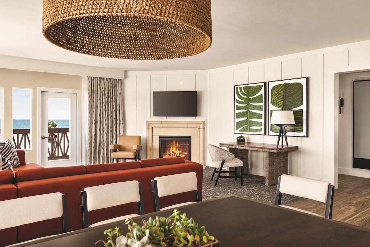 A modern living room with a fireplace, a large sofa, a dining table, and a view of the sea through the windows.