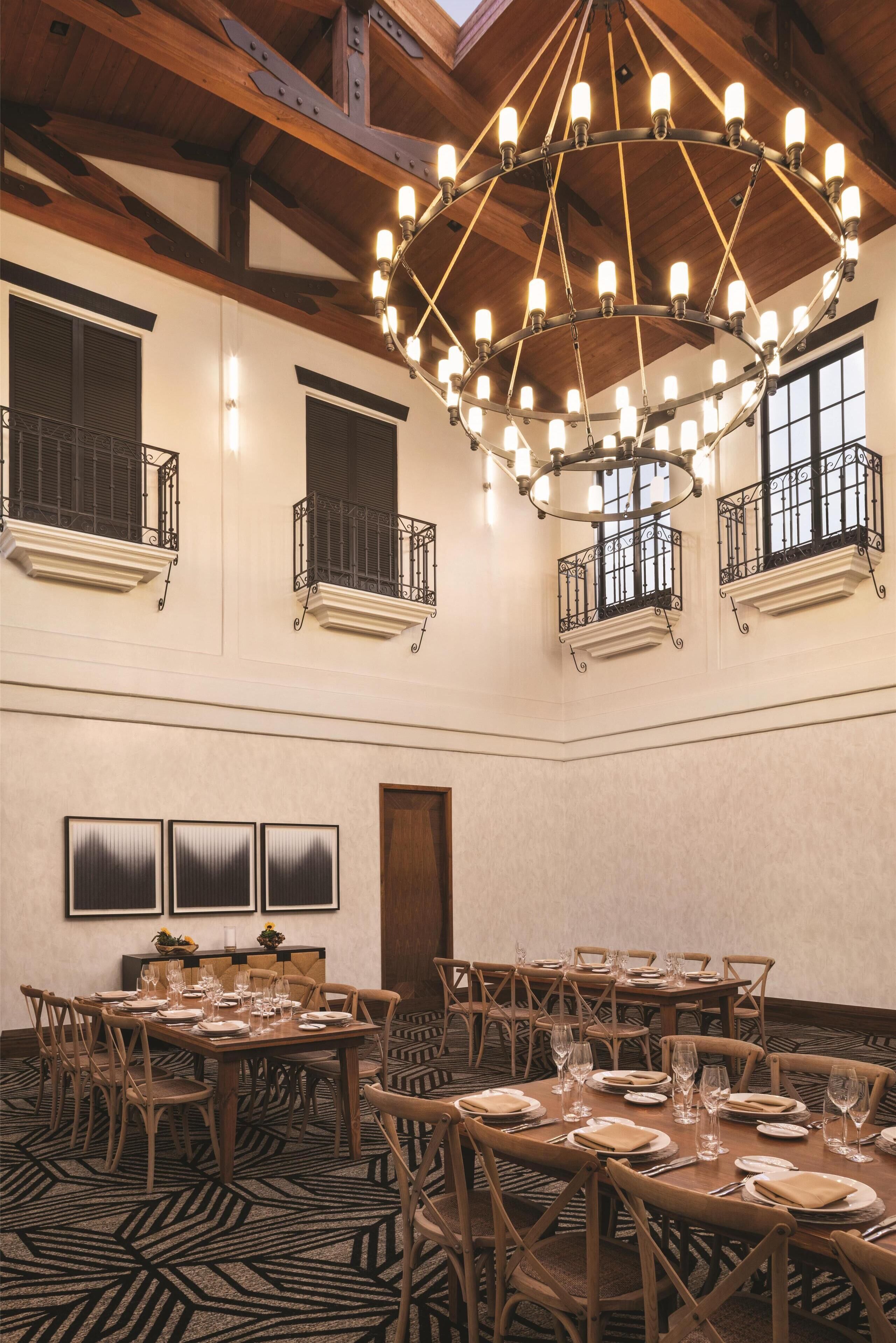 A spacious dining area with multiple tables set for a meal, a large chandelier, and a high ceiling with exposed beams.