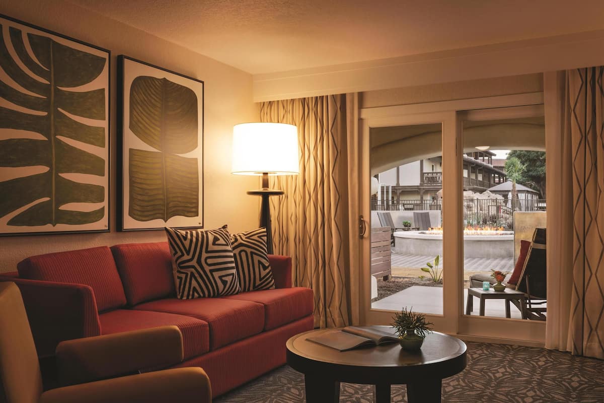 A hotel room with a red sofa, a round table with a book and a plant, and a large window with a view of an outdoor patio.