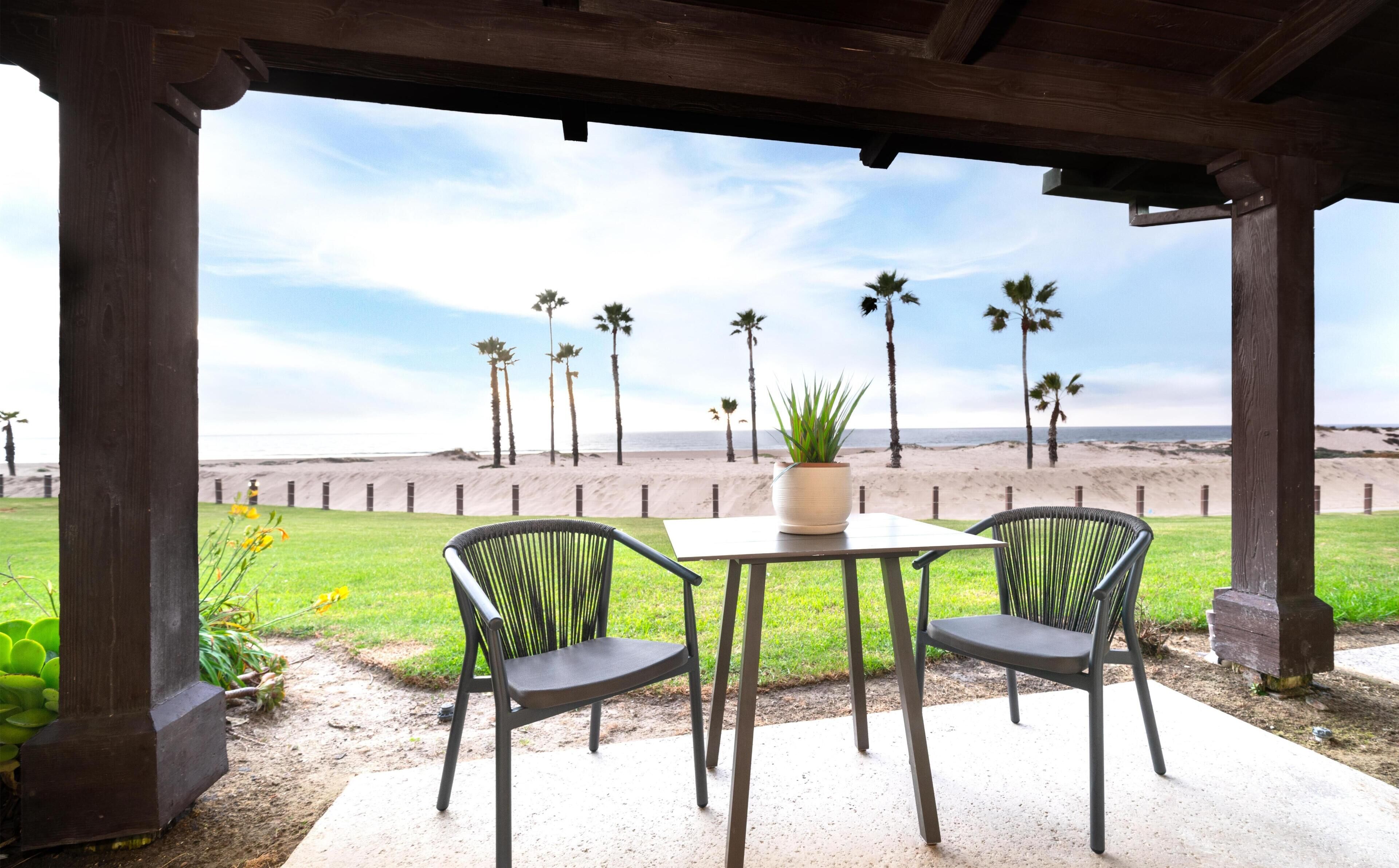 Un porche techado con dos sillas y una mesa, con vistas a una playa con palmeras y una orilla arenosa.