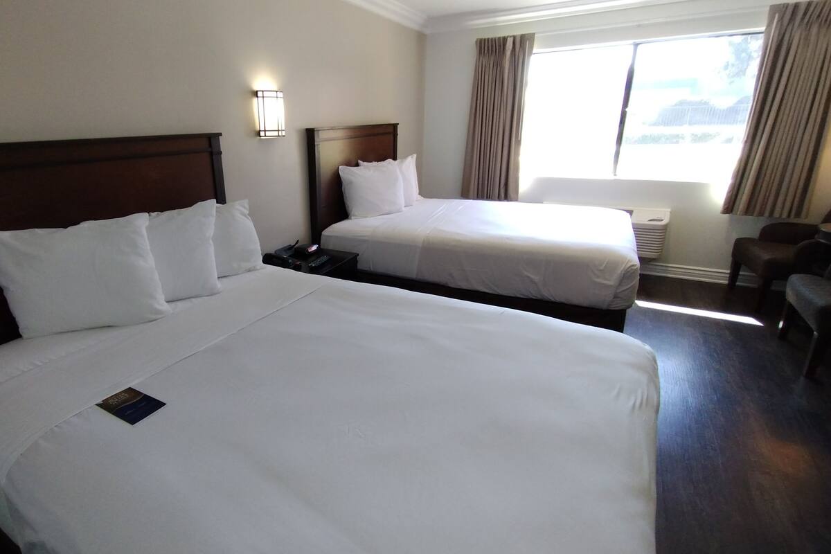 Ein Hotelzimmer mit zwei Einzelbetten, einem Nachttisch mit Telefon und Fernbedienung, einer Wandleuchte und einem Fenster mit Vorhängen.
