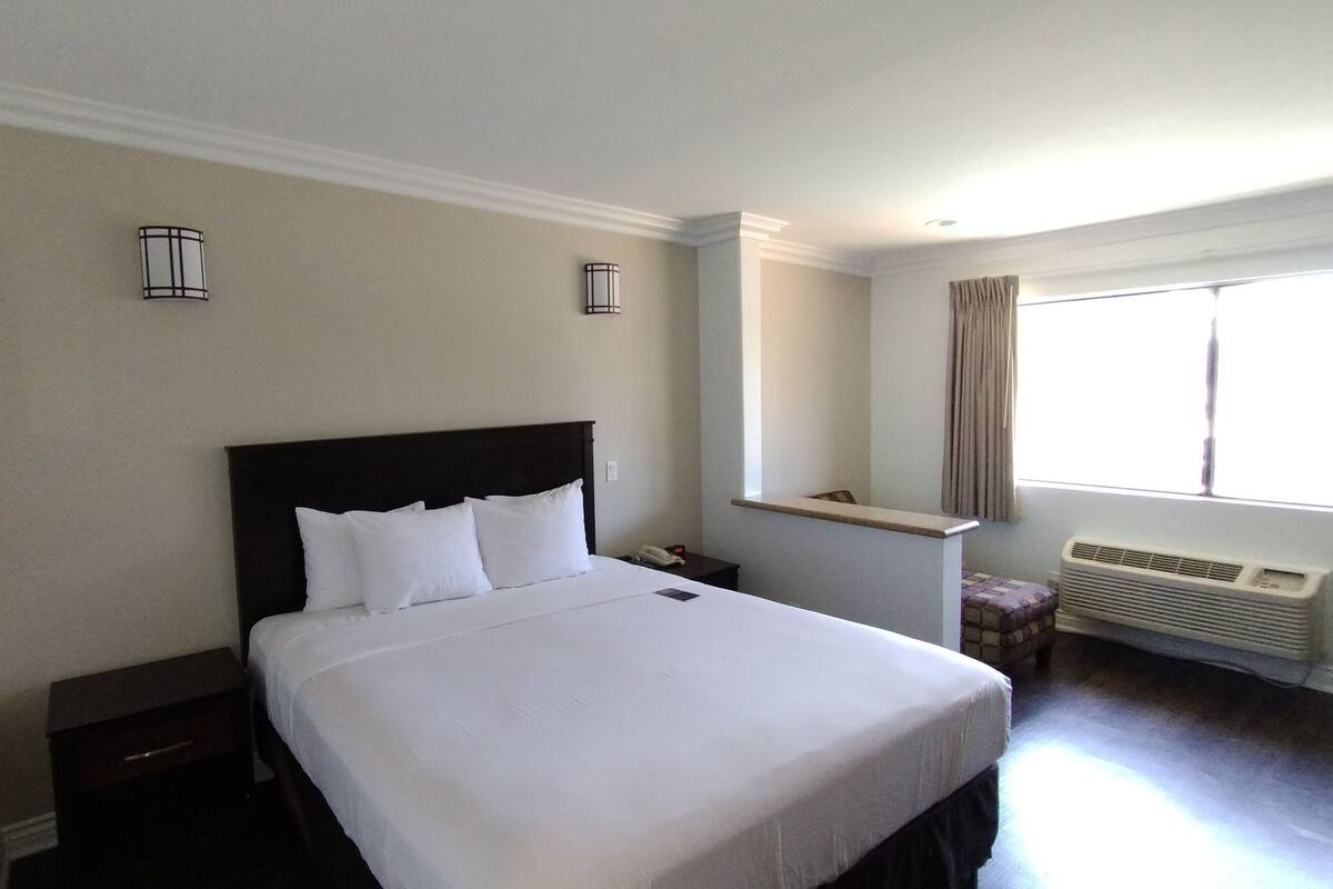 Ein Hotelzimmer mit einem großen Bett, einem Nachttisch, einer Wandlampe, einem Fenster mit Vorhängen und einer Klimaanlage.