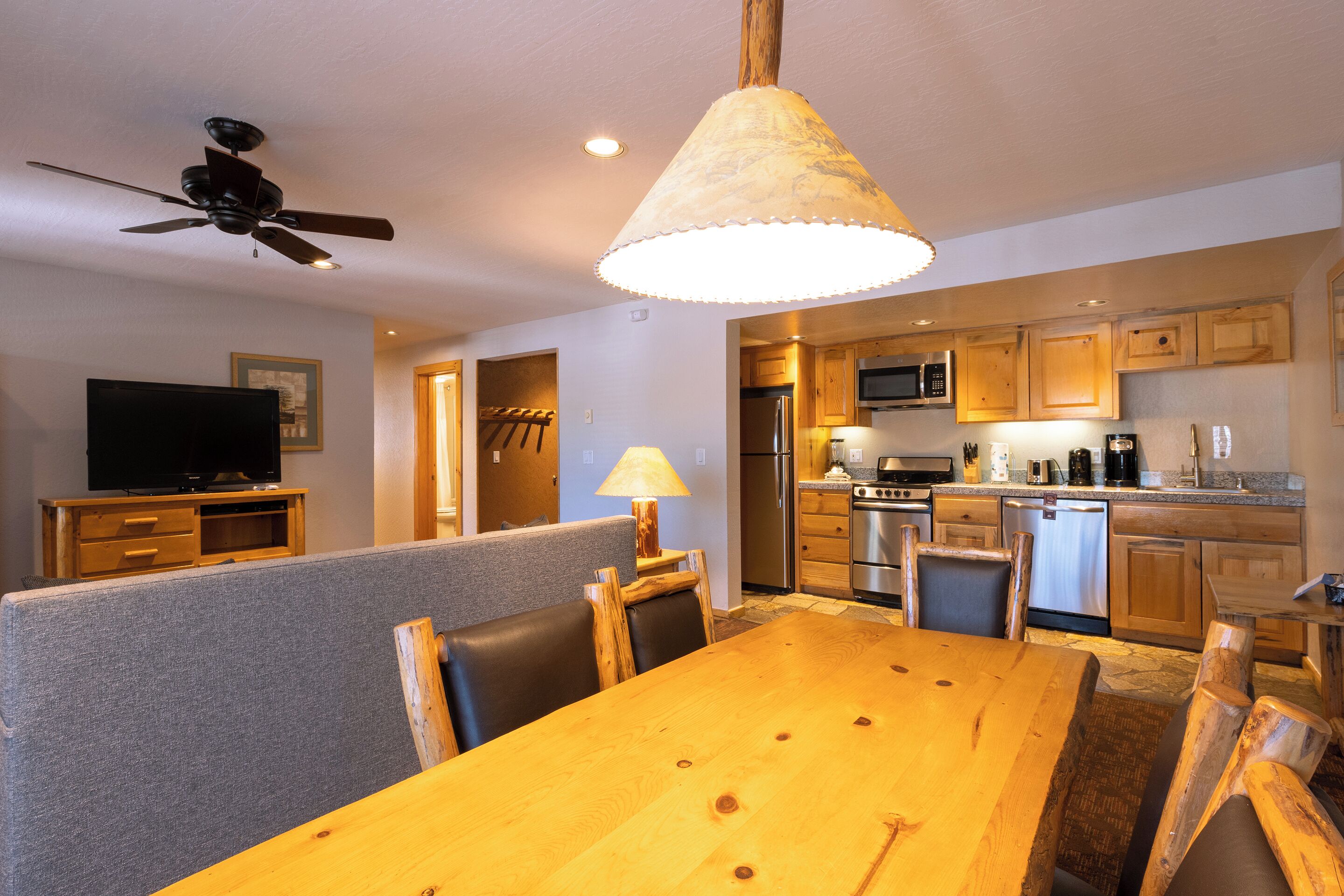 A dining area with a wooden table and chairs, a ceiling fan, a television, and a kitchen in the background.