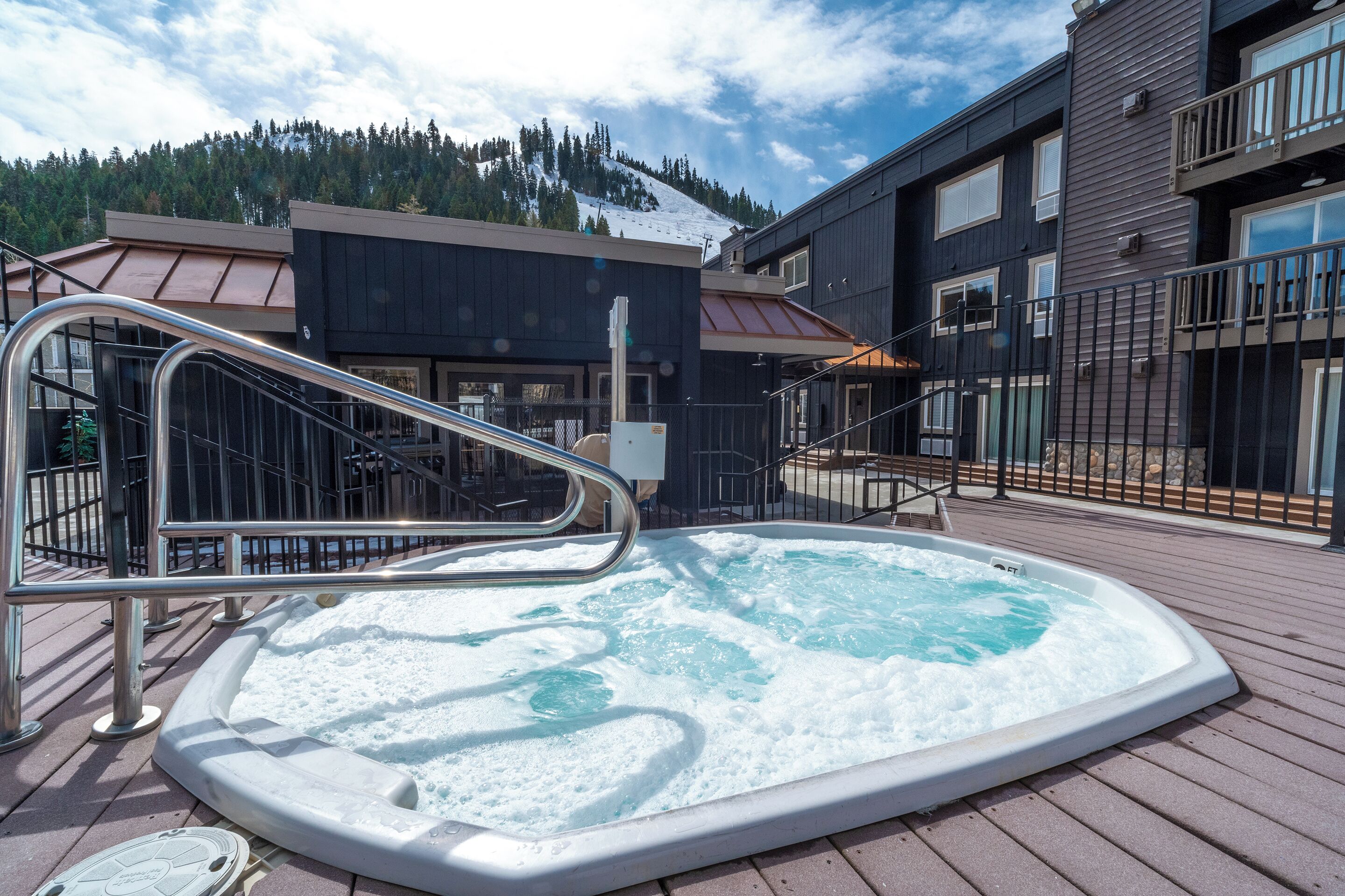 A hot tub on a wooden deck with metal railings, surrounded by buildings with large windows and a snowy mountain in the background.