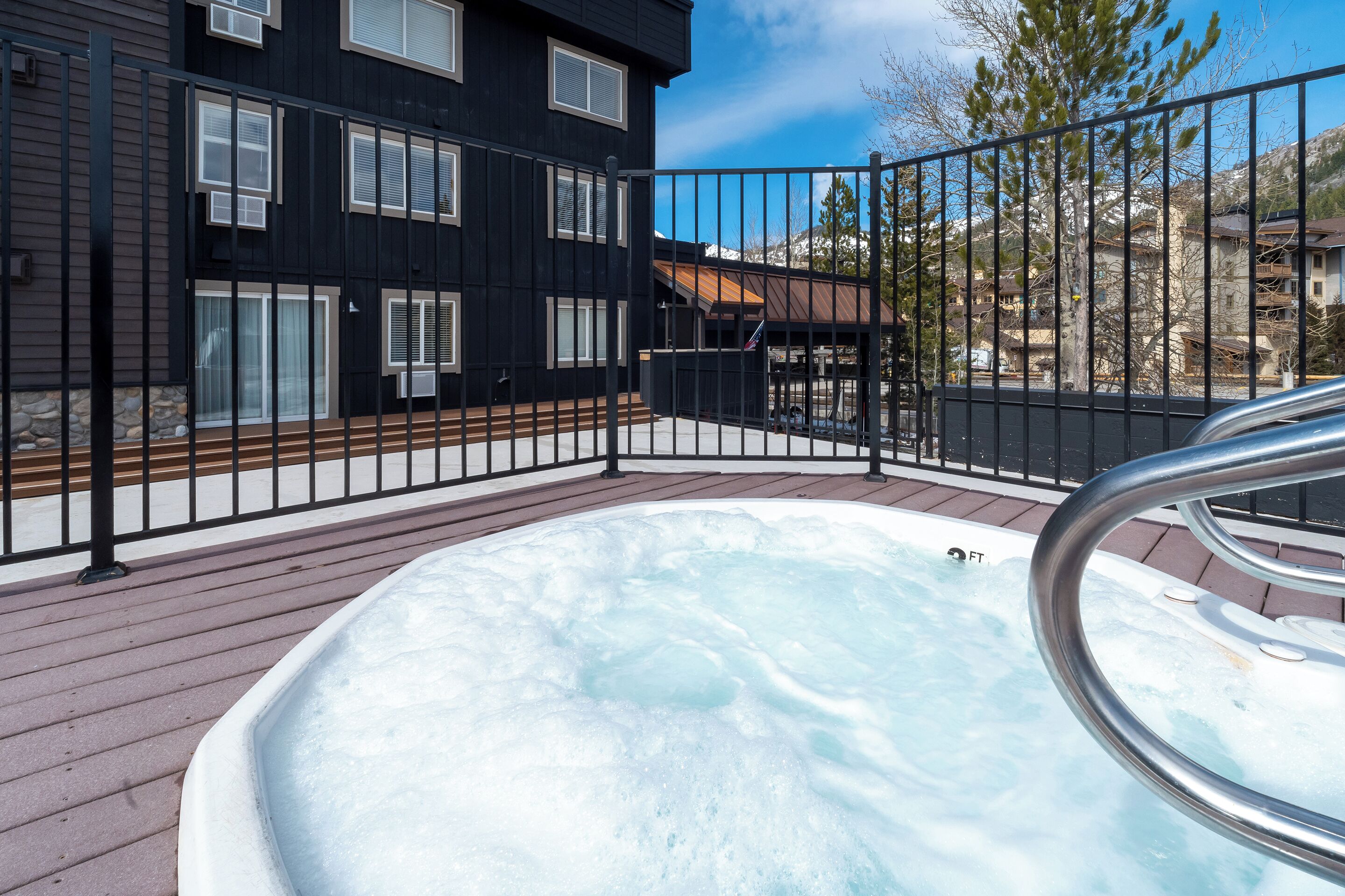 Un jacuzzi en una terraza de madera con barandilla metálica, rodeado por una cerca negra, y un edificio con amplias ventanas al fondo.