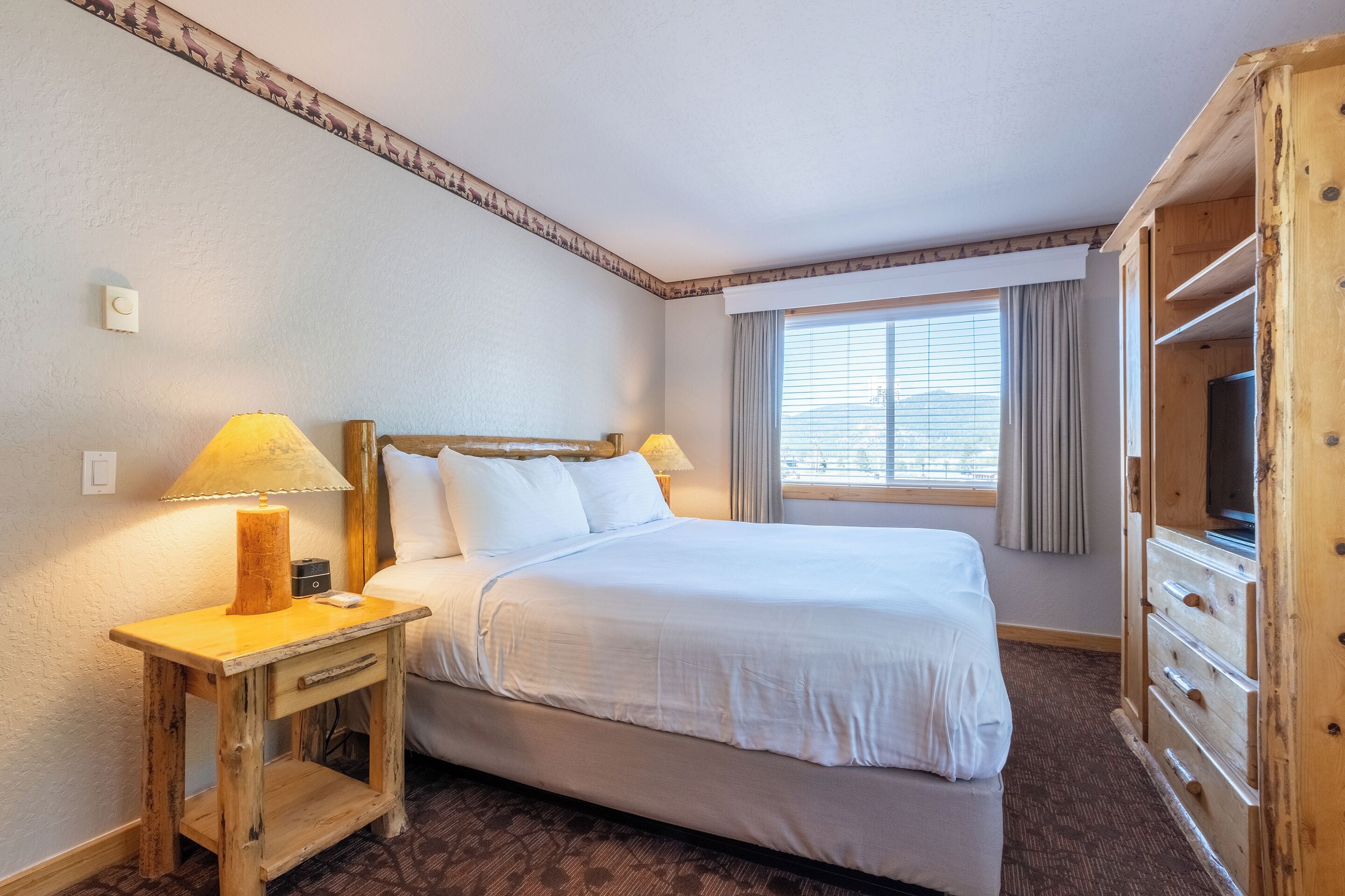 A hotel room with a large bed, wooden nightstands, a TV cabinet, and a window with curtains.
