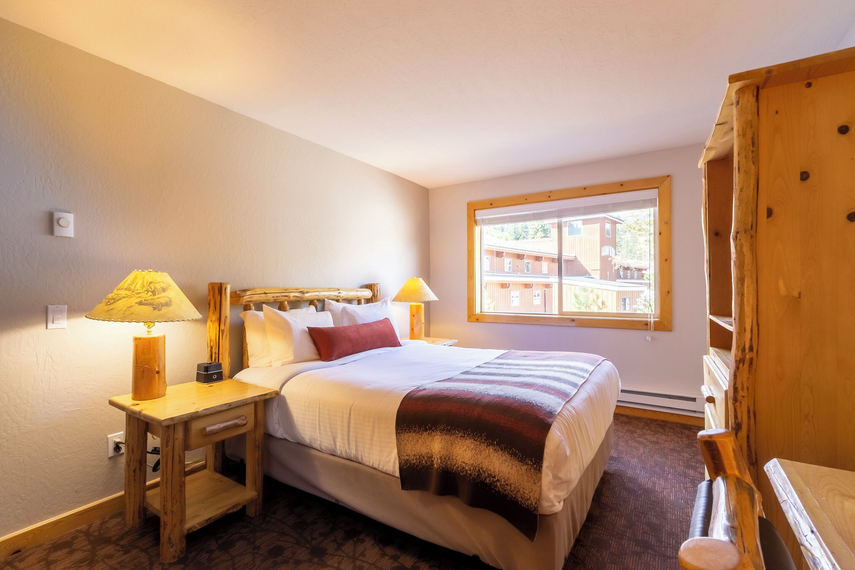 A neatly arranged bedroom with a bed, two bedside lamps, a window with a view, and a wooden wardrobe.