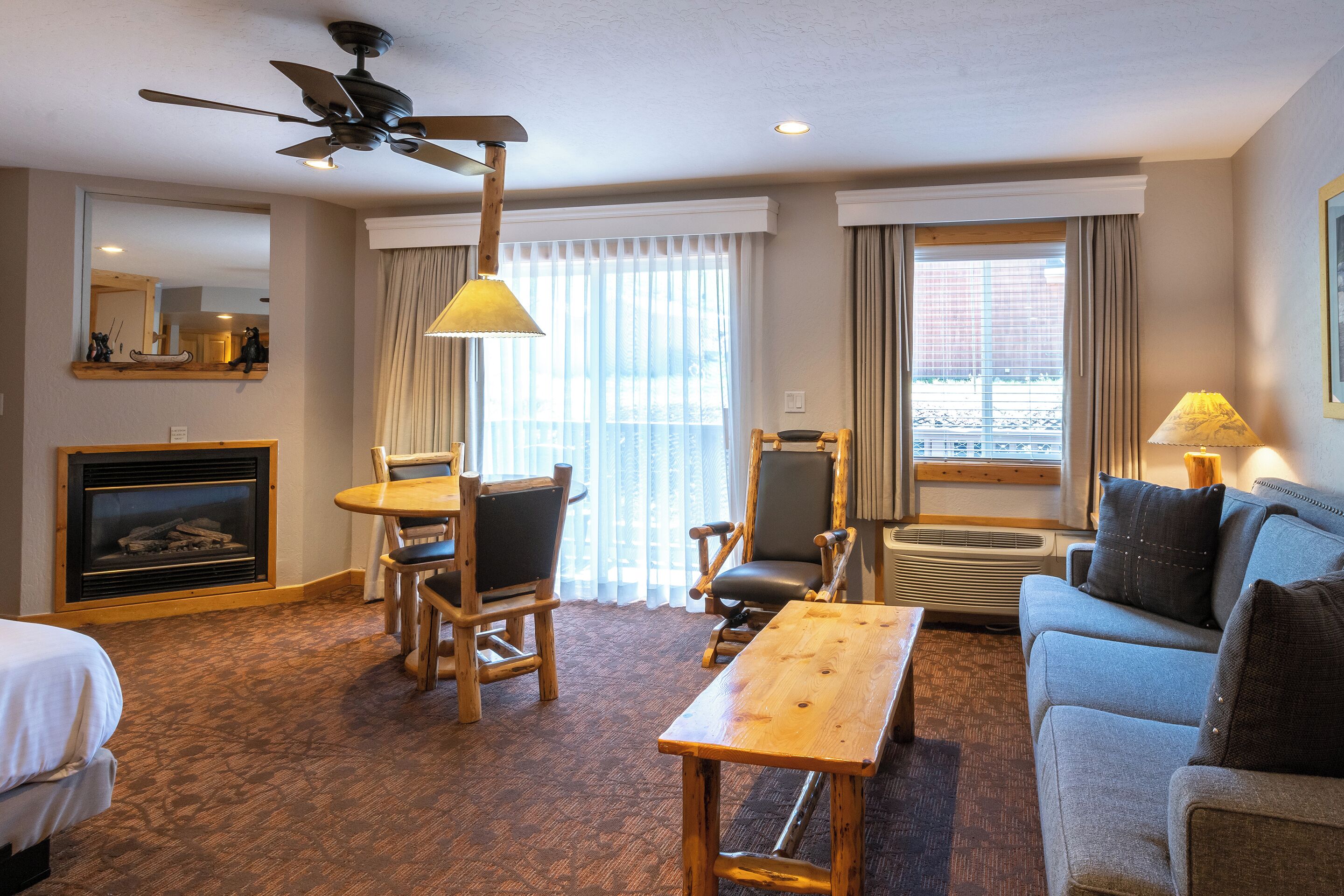 A hotel room with a wooden table, two chairs, a sofa, a ceiling fan, a fireplace, and large windows with curtains.