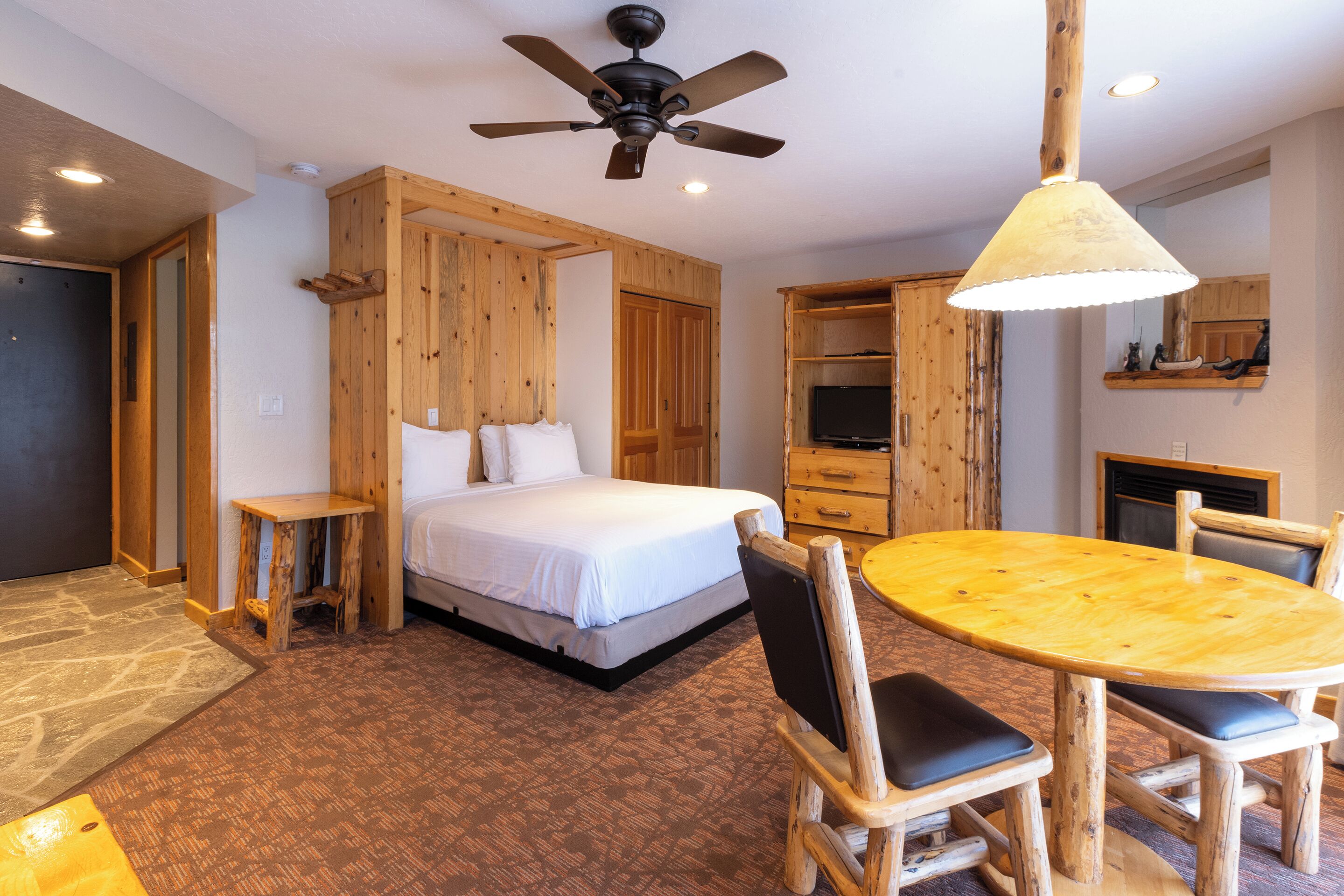 A bedroom with a bed, wooden walls, a ceiling fan, a wooden table with chairs, and a television.
