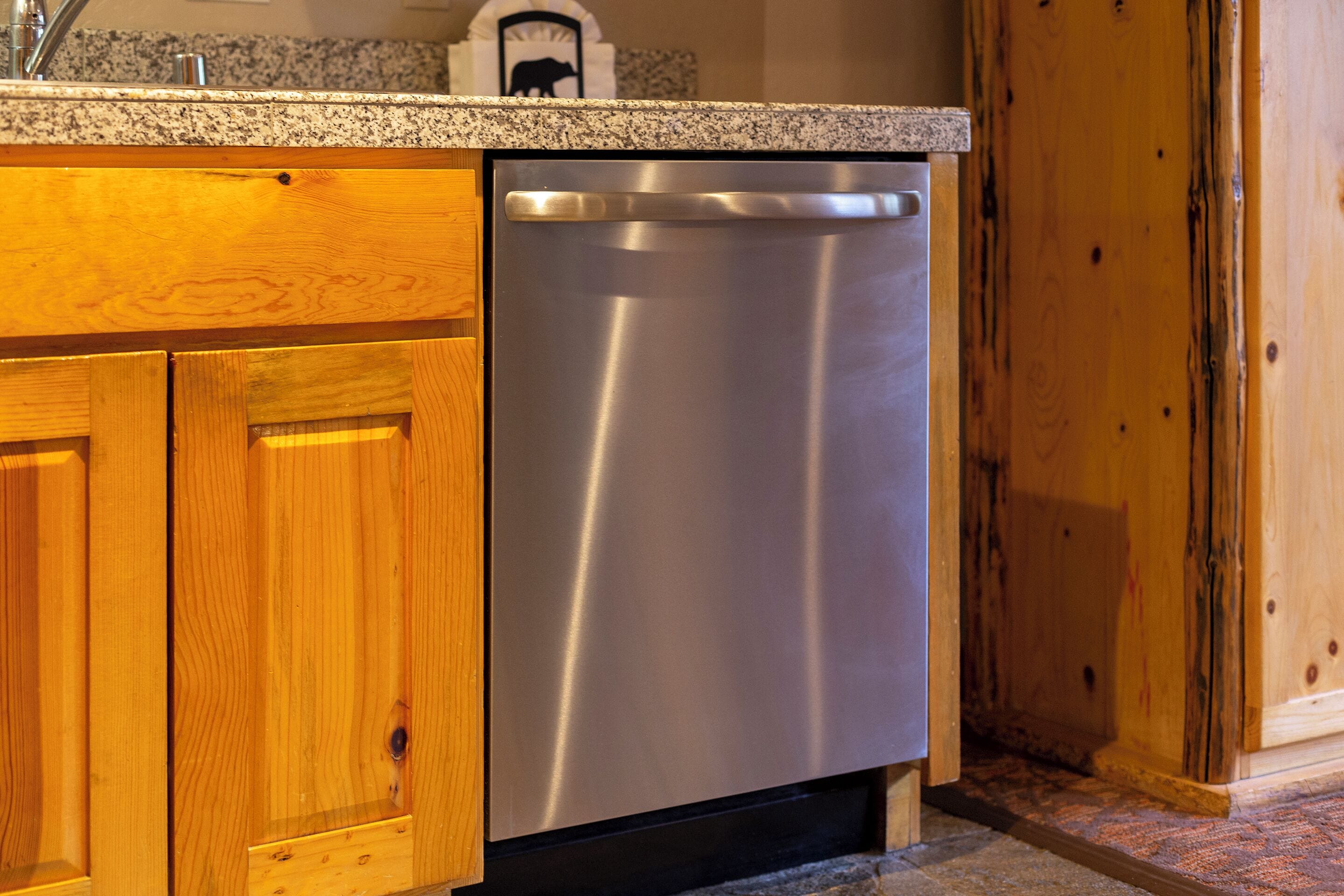 A stainless steel dishwasher installed under a granite countertop in a kitchen with wooden cabinets.