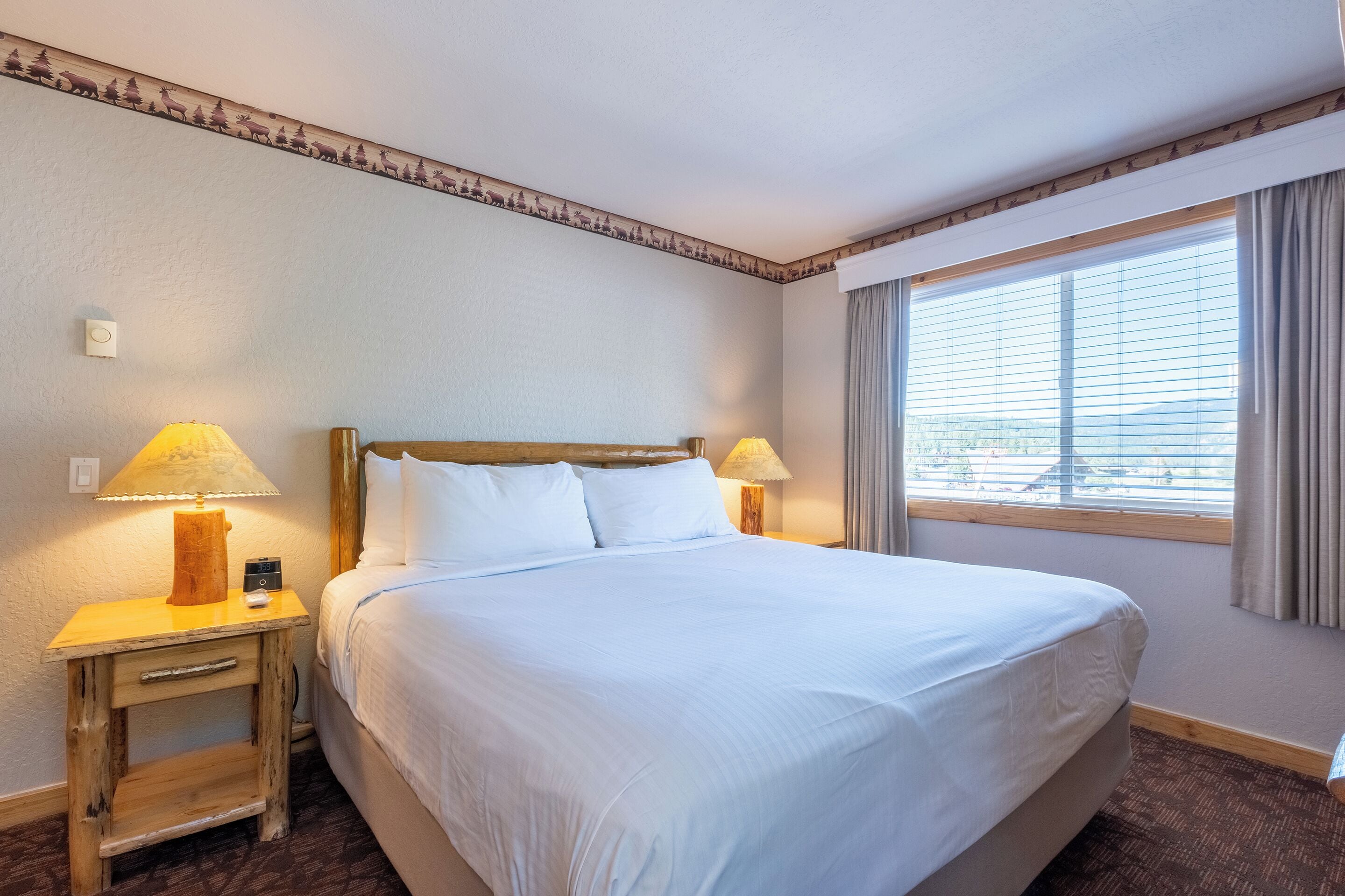 A hotel room with a large bed, two bedside lamps, a nightstand, a window with curtains, and a wall with a decorative border.