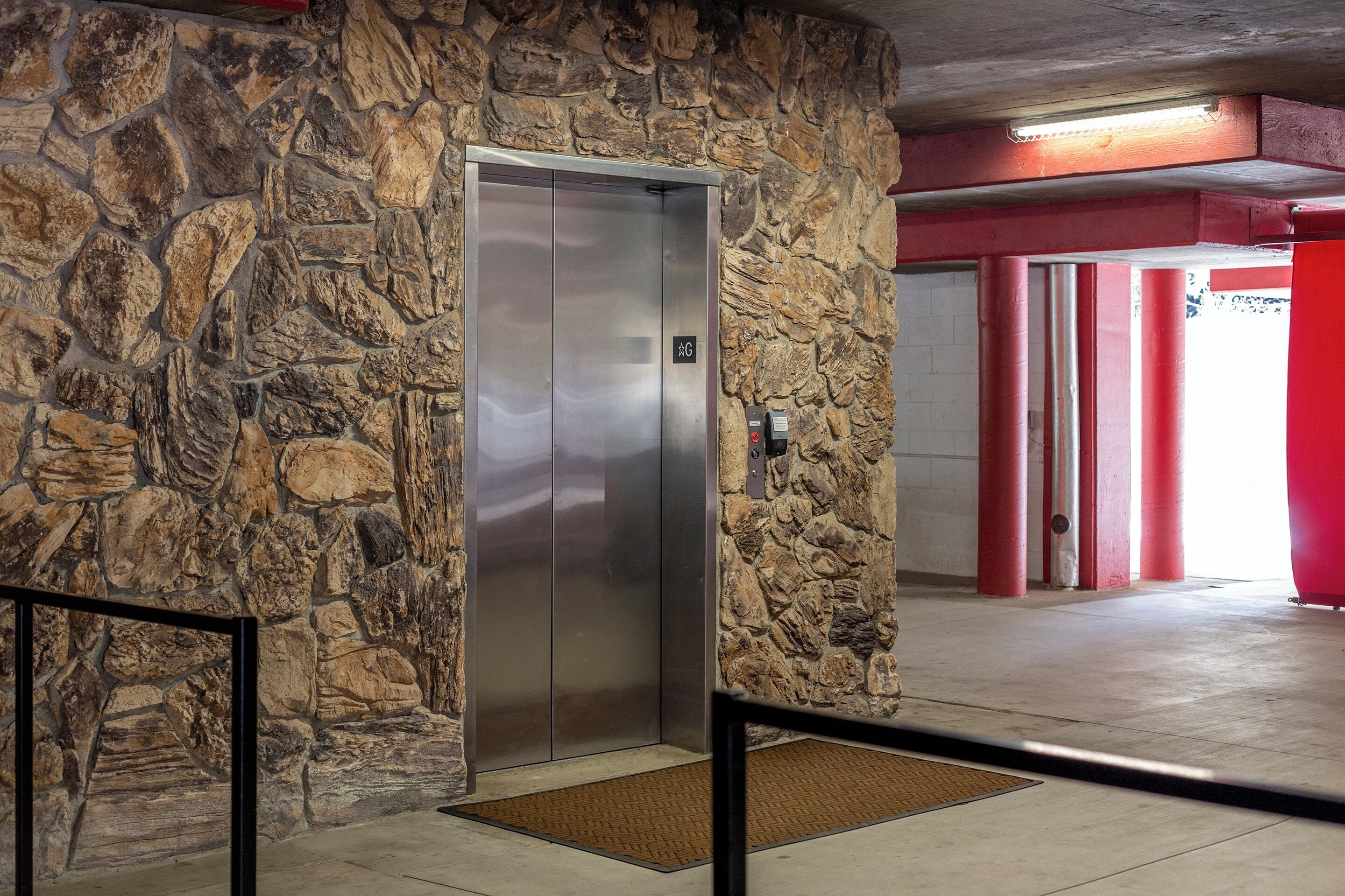An elevator with a stone wall, a red pillar, and a doormat on the floor.